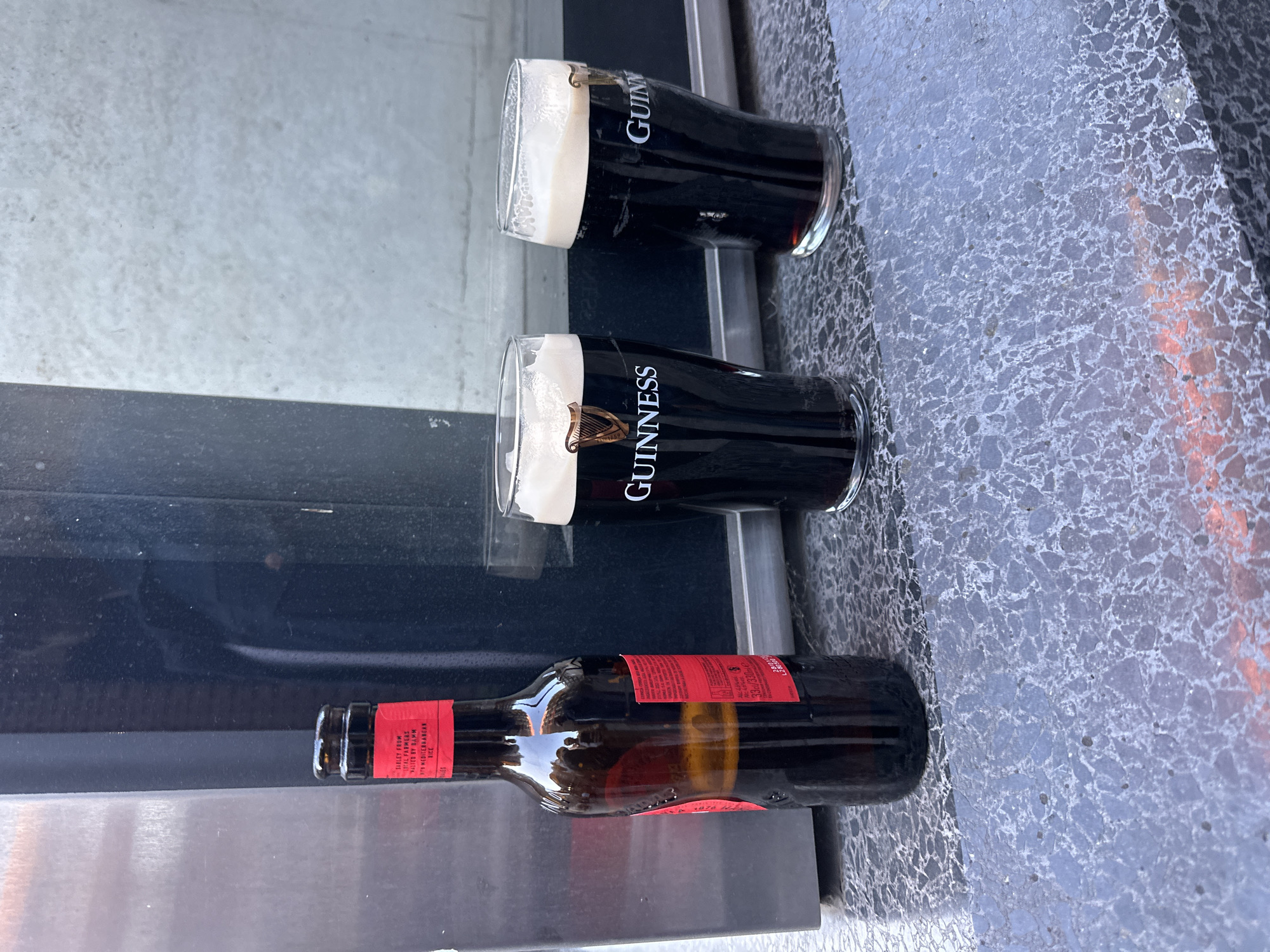 This media file captures an outdoor scene featuring three alcoholic beverages. The image is oriented sideways, with the contents appearing to defy gravity. When viewed upright, two full glasses of dark stout, identifiable as Guinness by the distinct harp logo and "GUINNESS" text on the side, are prominently displayed. Each glass has a thick, creamy white head.
To the right of the glasses, a dark glass bottle with a red label rests horizontally. Visible text on the red label includes "BARLEY FROM", "SITLEY FARMERS", "TED BY BURNS", "VIA MEDITERRANEAN", "33 cl", "330ml", "ALE", and partially "No. 1876". The bottle's contents appear dark, consistent with a stout or dark ale.
The drinks are situated on a speckled, light gray surface, possibly a stone or granite countertop or table, which shows some reflective qualities. A dark, reflective surface runs parallel to the drinks, perhaps a window or another part of the table. In the background, a lighter, possibly off-white or gray wall is visible.
The bright lighting suggests it is daytime. The setting appears to be an outdoor area, possibly a pub patio or cafe in London, United Kingdom, although no specific landmarks are visible to confirm the exact location beyond the provided context. No people are visible in the frame, and there is no explicit activity occurring beyond the presence of the drinks.