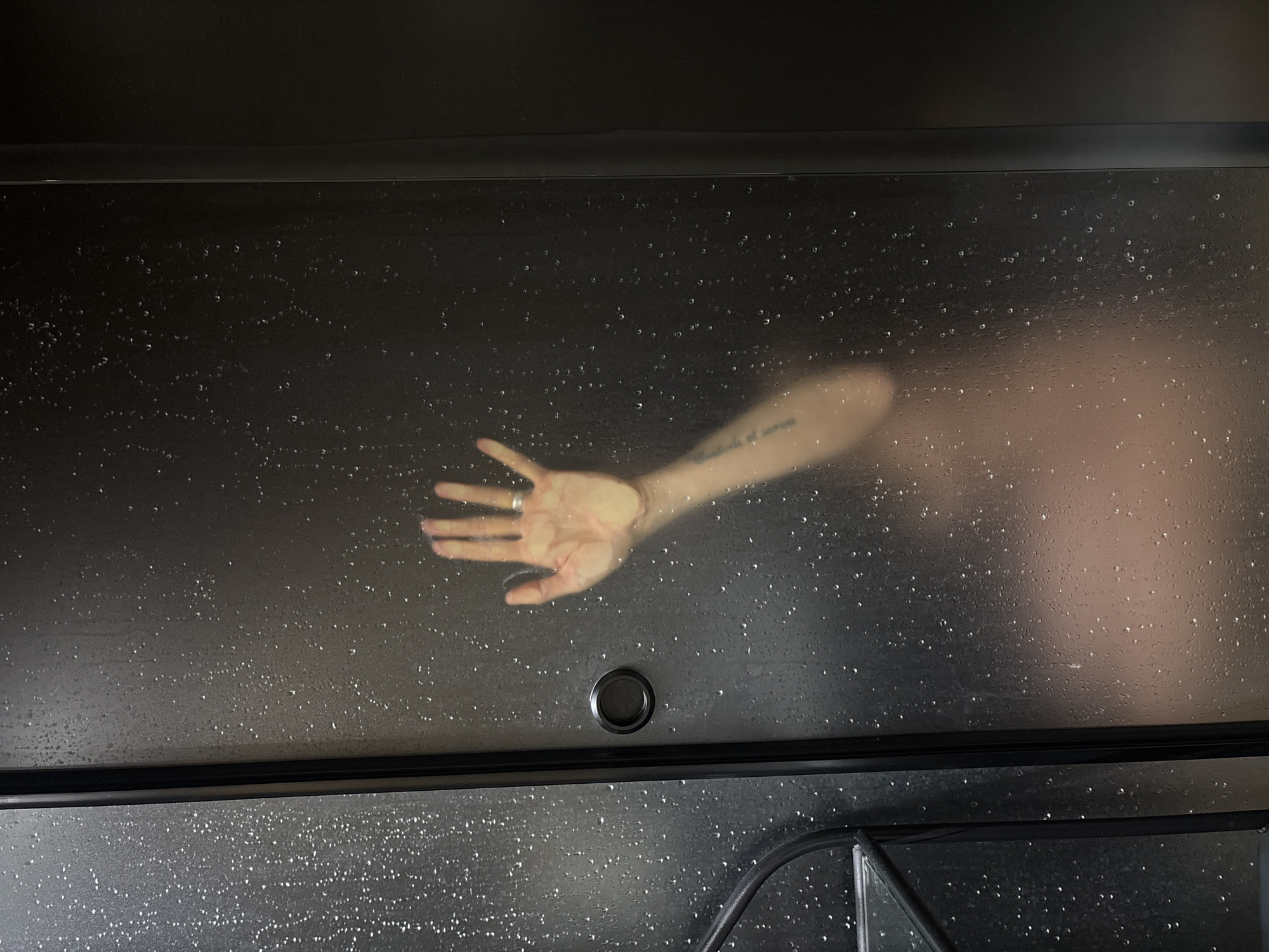 The image shows a person's hand and arm pressed against a dark, frosted glass surface. Water droplets are visible on the glass. A black frame surrounds the glass panel, suggesting it might be a shower door. A round, black knob is located on the glass panel. The tattoo is visible on the arm. There is not sufficient information to determine the location of the city of Chaikovsky, Russia.