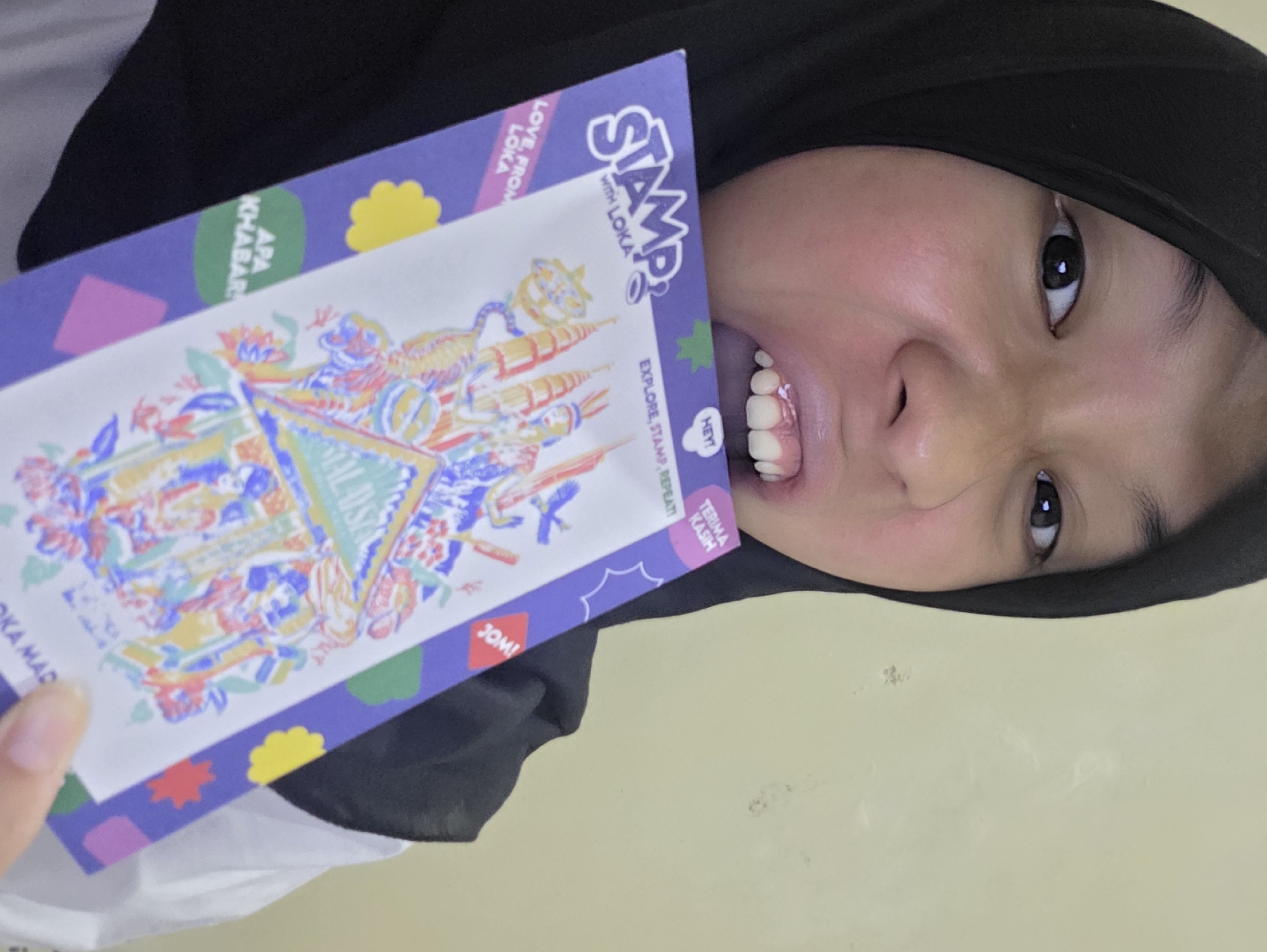 A person wearing a black head covering is holding a colorful item with the word "STAMP" on it. The background appears to be a plain wall. The text on the item also includes the phrases "WITH LOKA", "EXPLORE, STAMP, REPEAT!", "APA KHABAR", and "TERIMA KASIH". The image is taken at an angle, partially showing the person's face.
