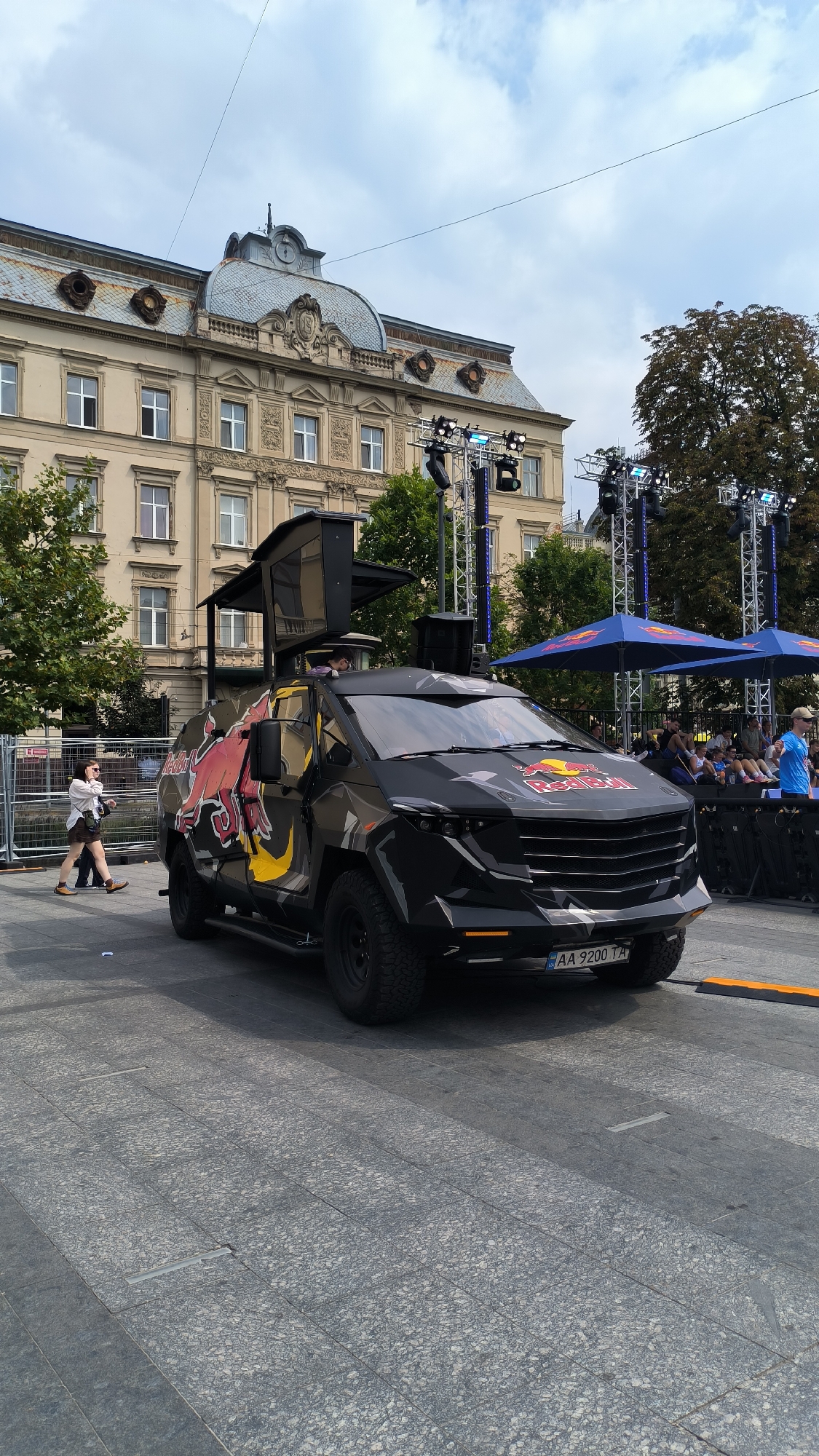 The image captures an outdoor scene in Lviv, Ukraine, likely during an event. In the foreground, a black, rugged vehicle with the Red Bull logo prominently displayed on its side and hood is parked on a paved surface. The vehicle has an open rear section, revealing what appears to be speakers or equipment. A person is visible standing in the open rear of the vehicle. To the left of the vehicle, a woman in a white top and brown shorts is walking away from the camera, holding a bag. Behind the vehicle, a stage with lighting equipment and large blue umbrellas with the Red Bull logo is set up. In the background, a multi-story building with a distinctive architectural style, featuring a mansard roof and ornate facade, stands under a cloudy sky. Several trees are also visible, along with a crowd of people gathered behind a barrier near the stage. The license plate of the vehicle reads "AA 9200 TA."