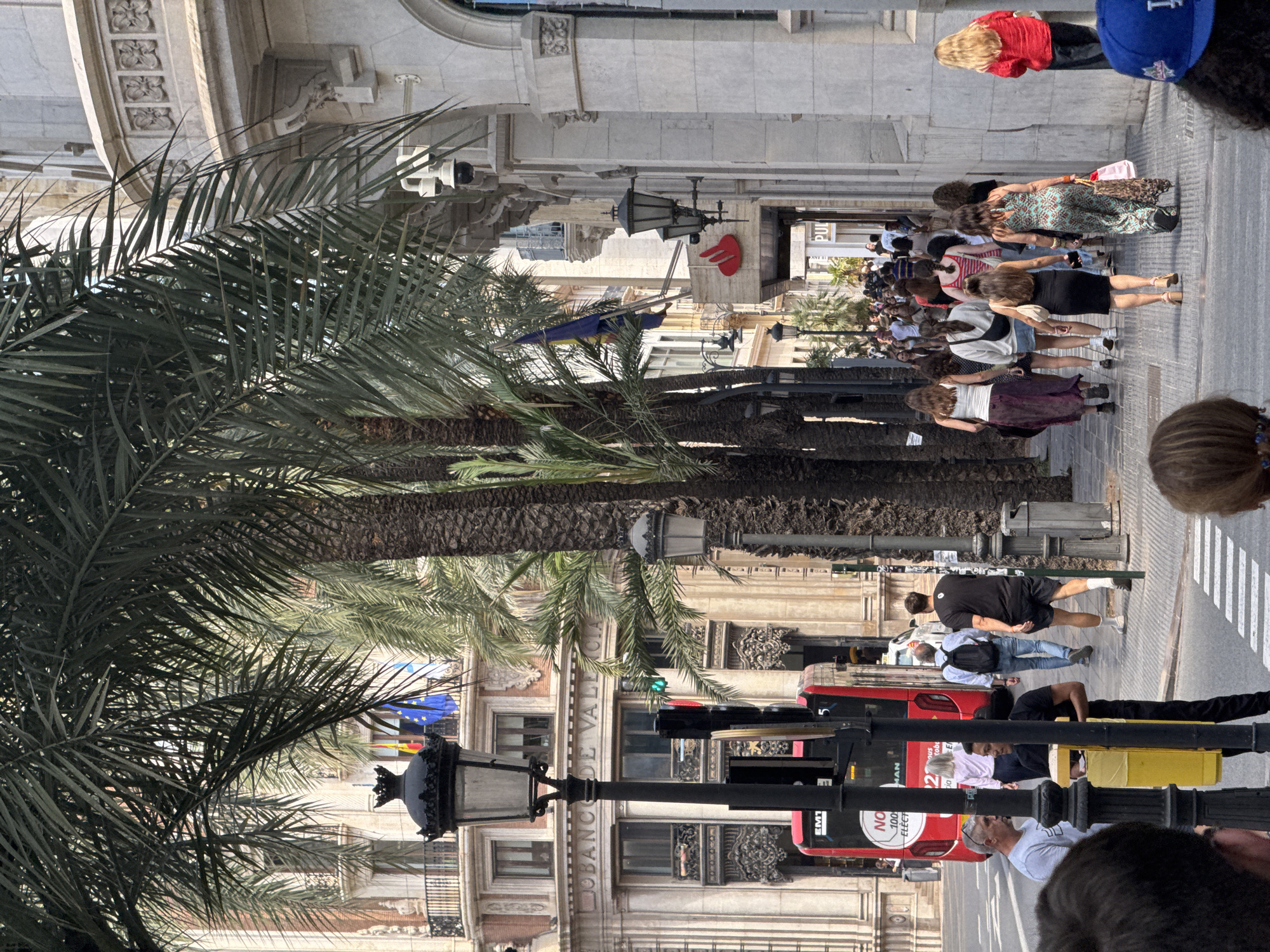 In Valencia, Spain, a bustling street scene unfolds beneath the shade of tall palm trees. On the left, the ornate facade of the "BANCO DE VALENCIA" building stands prominently, adorned with flags. A red public transport bus is stopped, its "EM1" designation visible. A traffic light shows red and green signals. Pedestrians walk along the sidewalk on both sides of the street.
On the right side of the street, a white building features architectural details and multiple security cameras. A red "SANTANDER" logo is visible on a storefront. The sidewalk is paved with rectangular tiles. In the foreground, several people are walking away from the camera, some carrying shopping bags. The overall impression is a lively urban environment in Spain.