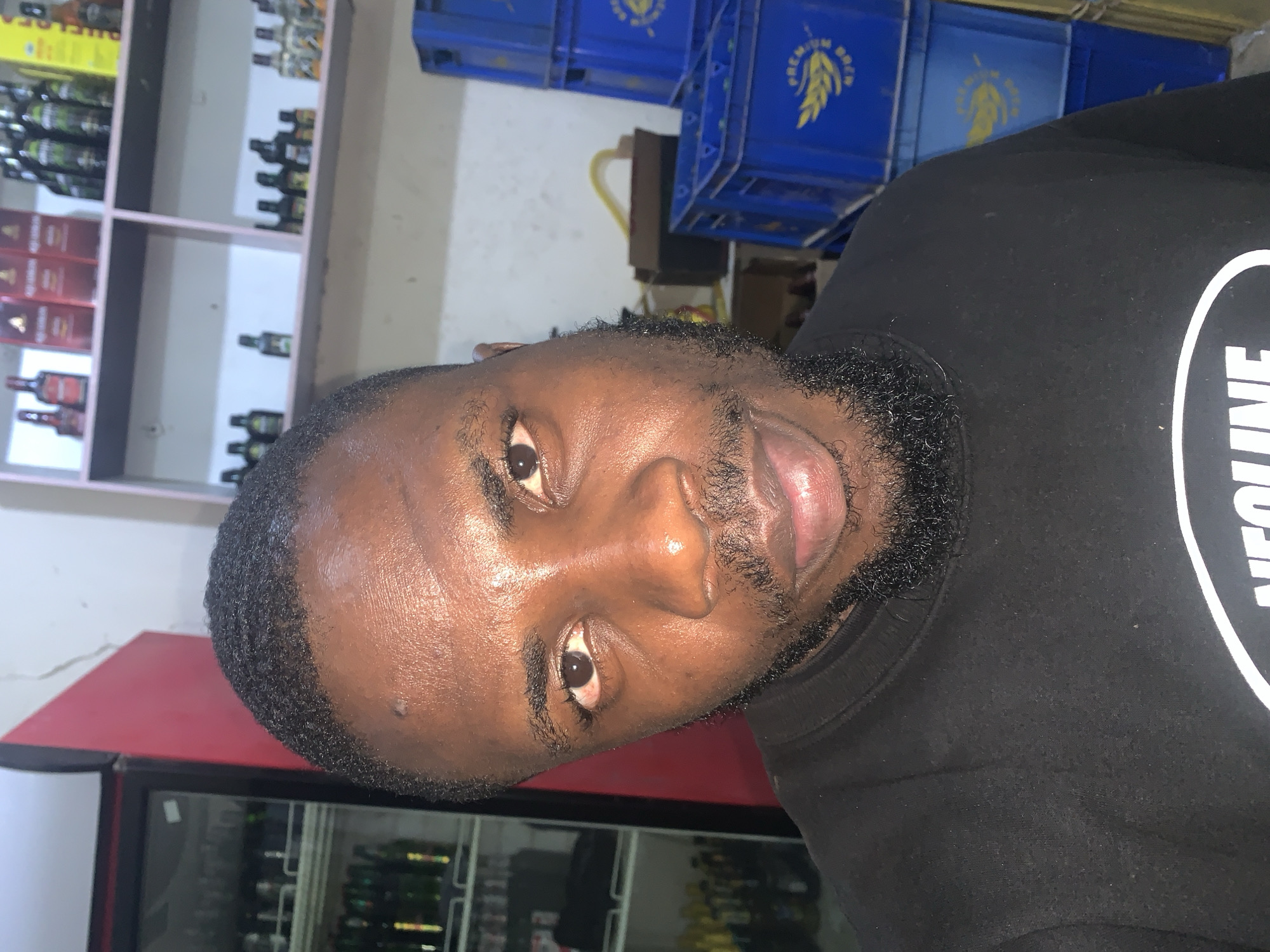 A medium shot captures a Black male in his late 20s or early 30s, facing the camera from a slightly low angle. He has short, wavy black hair and a neatly trimmed black beard and mustache. His skin is dark brown and appears slightly dewy, possibly from sweat. He is wearing a black t-shirt with a white logo on the chest, partially visible.
The background suggests a retail environment, likely a store or a warehouse. To the left, a red-and-black refrigerator with glass doors is filled with bottles, some of which are green. To the right, several stacked blue plastic crates are visible. The shelves behind him are stocked with various bottles, some appearing to be liquor or beverages, in a range of colors including amber, green, and red. The overall lighting is somewhat dim, with some bright spots illuminating the products on the shelves. The setting is identified as New Stadium, Nigeria.
