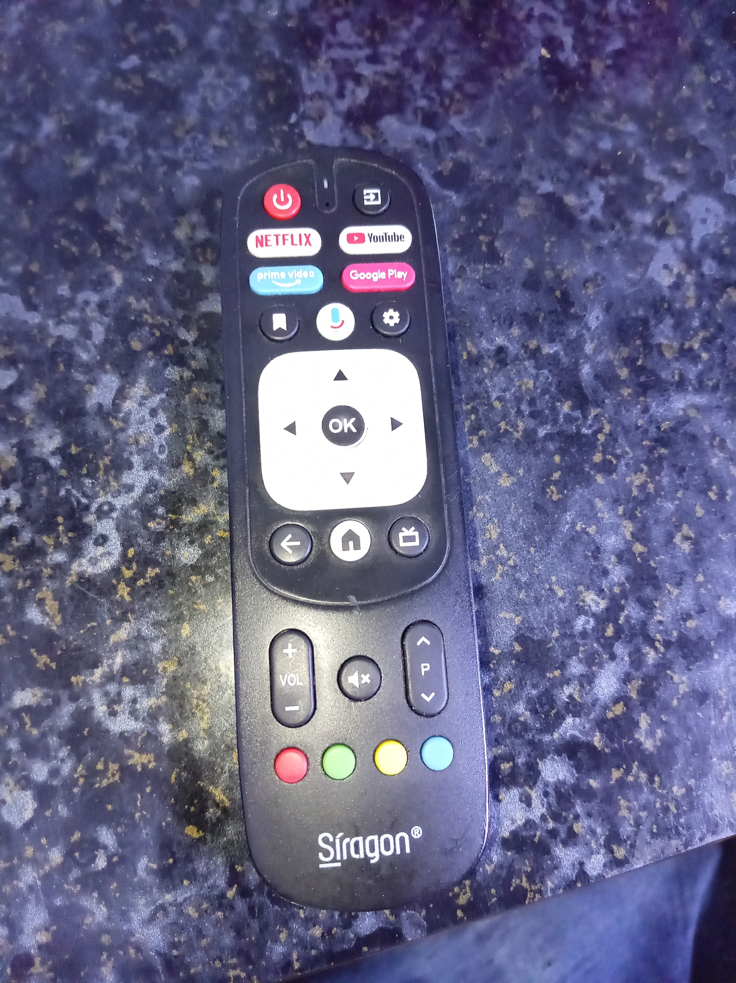 A close-up shot reveals a black television remote control resting on a speckled surface with hints of blue and gold, possibly a countertop or table. The remote features buttons for power, Netflix, YouTube, Prime Video, Google Play, microphone, settings, directional navigation with an "OK" button, back, home, menu, volume control, mute, channel change, and four colored buttons (red, green, yellow, blue). The brand name "Stragon" is printed at the bottom of the remote. The image provides no further information to establish the location in Tinaquillo, Venezuela. No people, subjects, or interactions are visible.