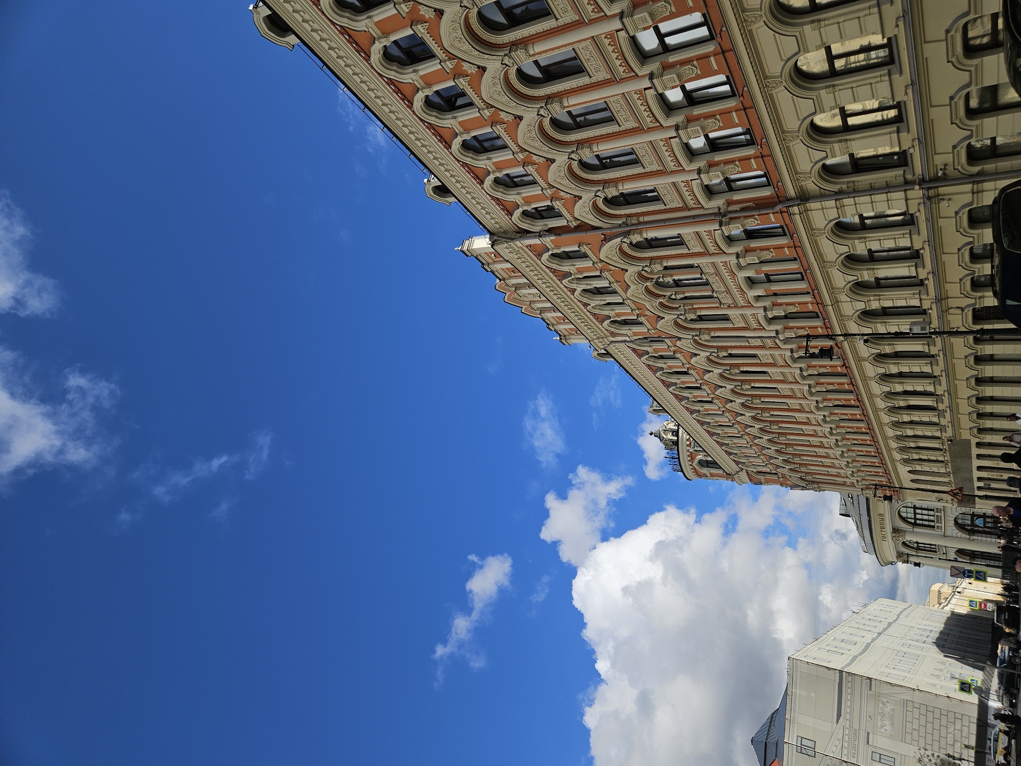 The image shows a view from the ground looking up at a building on a street in Moscow, Russia. The building's facade is ornate, with orange and beige accents and many arched windows. The sky is blue with scattered white clouds, some near the top corner of the building and others closer to the bottom. People are visible walking on the sidewalk near the base of the building. Other buildings, street signs, and parked cars suggest a busy urban environment.