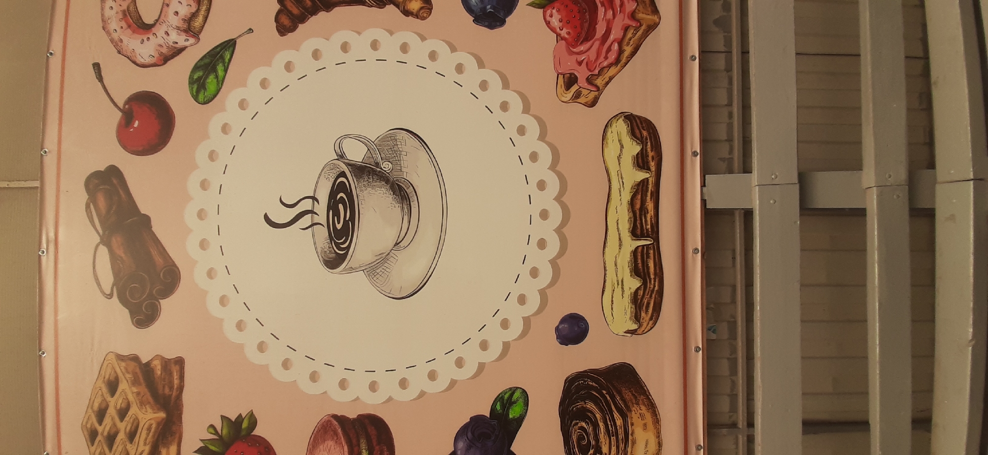 The image depicts a close-up view of a banner featuring various baked goods and a cup of coffee, in Yessentukskaya, Russia. The banner is hung outdoors, with metal fixtures visible along the top edge, suggesting it is part of a temporary display.
Dominating the central portion of the banner is a hand-drawn illustration of a steaming cup of coffee, placed within a circular, lace-like border. The coffee is depicted with swirling steam rising from it.
Surrounding the central coffee illustration are several other drawings of food items. In the upper left, there are stacked waffles and cinnamon sticks. To the right of these, a cherry and a pink frosted donut are visible. Below the coffee cup, on the left, are macarons and blueberries. Further down and to the left, a swirl-shaped pastry, possibly a cinnamon roll, is shown. On the right side, below the donut, is a croissant, followed by a long eclair with white icing. In the lower right, a waffle topped with pink frosting and a strawberry is depicted.
The background of the banner is a light pink color. The lower portion of the image shows the top of a grey, slatted fence or railing, with vertical wooden planks visible behind it, indicating the banner is displayed above this structure. The lighting suggests an outdoor environment, likely during daylight hours. There are no people or animals present in the image.