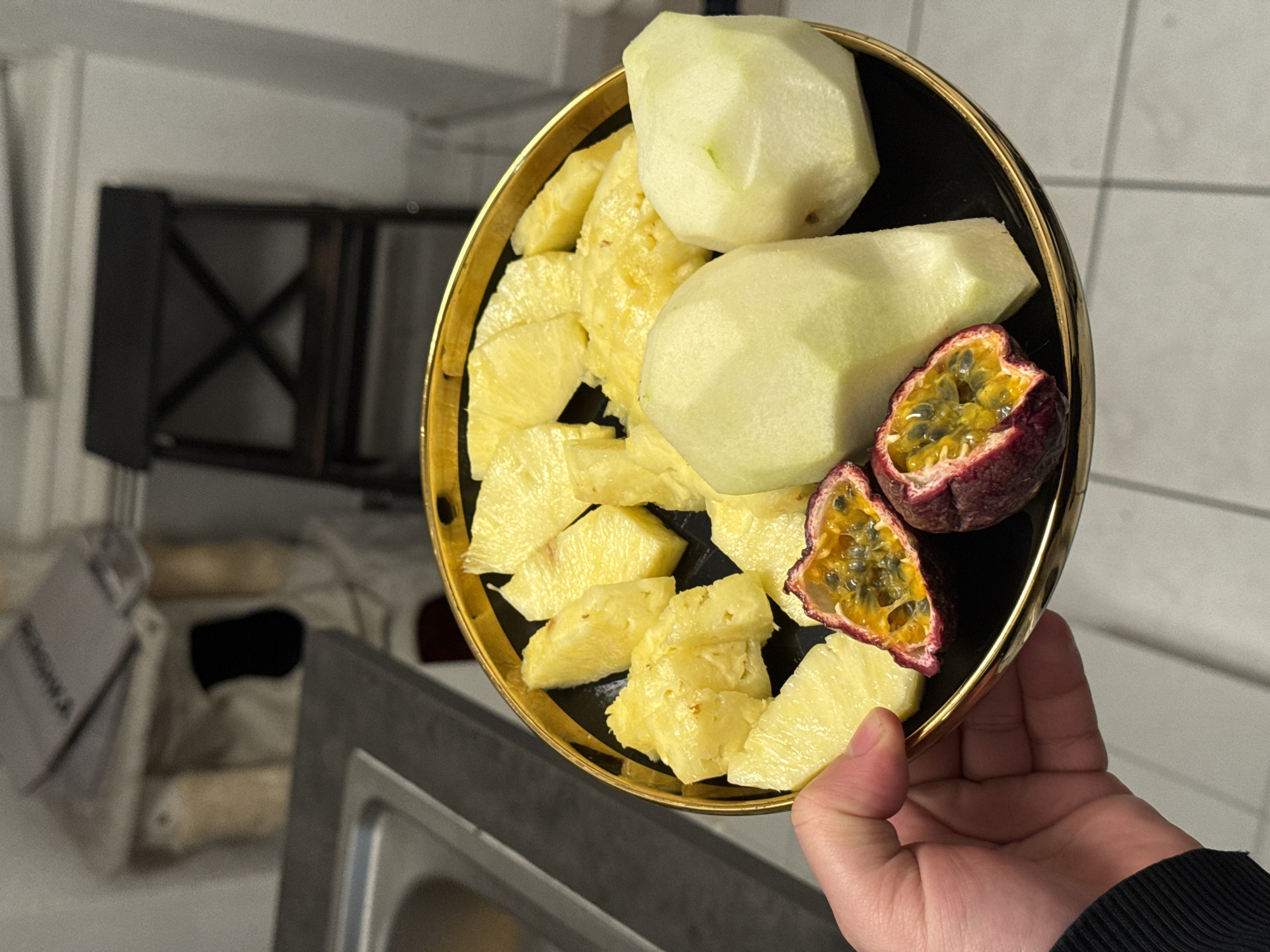 The scene is set within a domestic interior, likely a kitchen, in the city of Strasbourg, France. A human left hand, with light skin and visible knuckles, is prominently featured in the lower-left quadrant, holding a round, black plate with a distinct gold rim. The hand wears a black, ribbed long-sleeved garment.
The plate contains an assortment of prepared, cut fruit. Approximately two dozen irregular, yellow chunks of pineapple are arranged across the plate. To the right of the pineapple pieces are two larger, pale green-yellow, block-shaped pieces of peeled fruit, possibly melon or pear. In the lower-center of the plate, two halves of a dark reddish-purple passion fruit are visible, cut longitudinally to reveal their bright orange, gelatinous pulp filled with black seeds.
The background elements are blurred but discernible. To the left, a stainless steel sink basin is set into a dark gray countertop. Above and behind the sink, a light-colored wall features a cluttered shelf or storage area with indistinct items, including what appears to be stacked papers or magazines and some cylindrical objects. To the right, a dark wooden chair with an "X" shaped backrest is partially visible against a plain white wall. The floor beneath the plate is light gray tiled, with visible grout lines forming a grid pattern.
