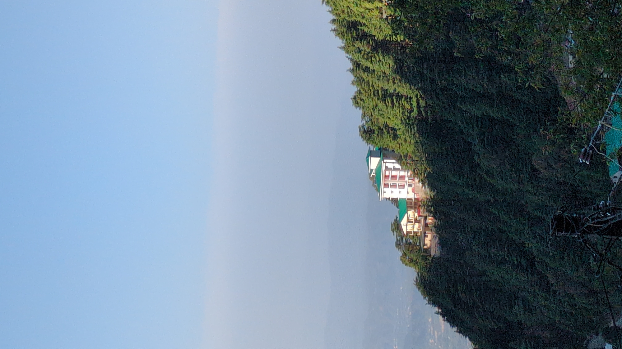 The image shows a building complex situated on a hillside in Haini, India. The buildings are white with red accents and green roofs, surrounded by dense green trees. The sky is a light blue with a hint of haze. Some power lines are visible in the lower right corner of the frame. The ground below the hillside is partially obscured by the haze.