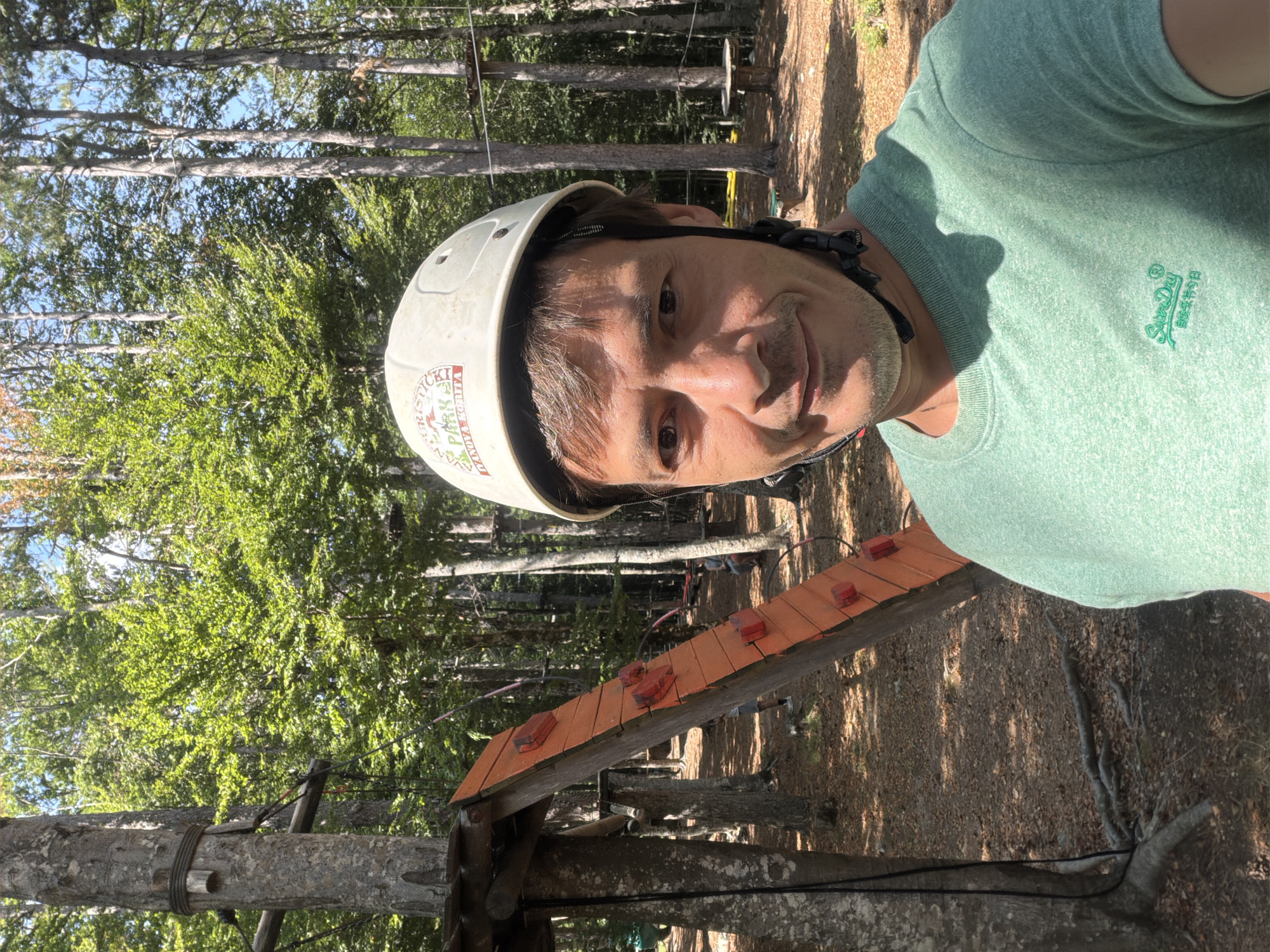 In an outdoor adventure park, a man is taking a selfie. He is wearing a white helmet with a logo that reads "PARK". He has short brown hair, brown eyes, and a light mustache. He is wearing a green t-shirt with a Superdry logo. The background shows a forest with tall trees, some of which have ropes and platforms for activities. There is an orange climbing wall with red grips leaning against a tree. The ground is covered in fallen leaves. The location is identified as Dolovi, Montenegro.