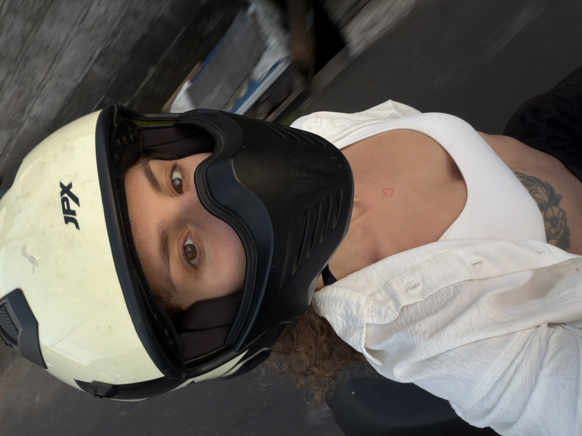 An overhead, slightly angled close-up image depicts a female individual in the city of North Kuta, Indonesia. She is wearing a cream-colored JPX brand full-face helmet with a black chin mask, partially obscuring her face except for her right eye and upper forehead. Her visible eye is brown. Curly, brown hair is visible beneath the helmet. She is dressed in a white bralette or crop top, overlaid by an unbuttoned white, textured long-sleeved shirt. A small red heart tattoo is present on her upper left chest, and a larger, dark tattoo, possibly a butterfly or floral design, is visible on her lower abdomen. The background is blurred, showing indistinct dark and light elements, suggesting an outdoor urban environment, possibly in motion.
