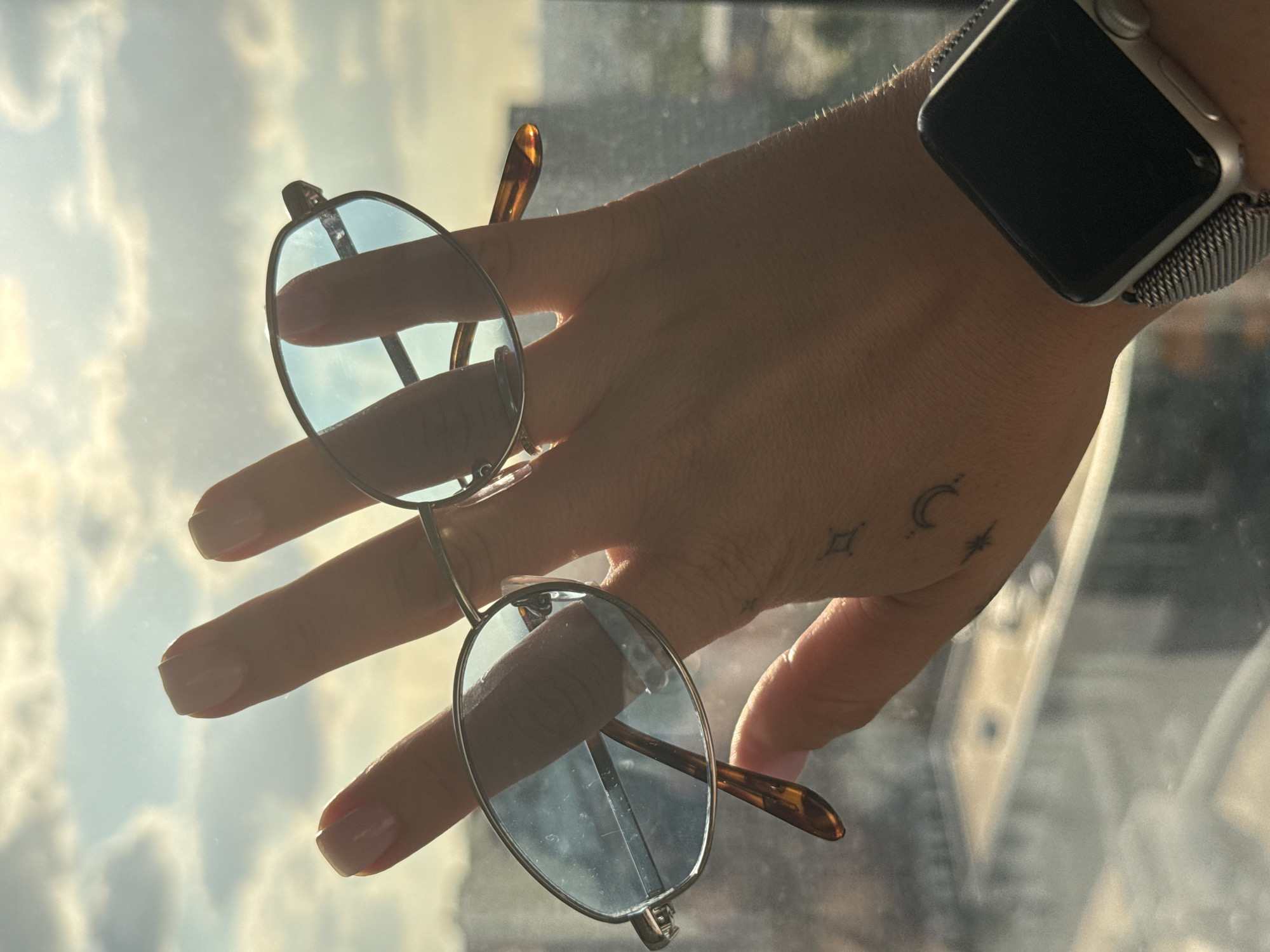 A close-up, medium shot captures a left hand, adorned with manicured nails and subtle tattoos of a crescent moon, a star, and a diamond shape on the wrist, holding a pair of wire-rimmed eyeglasses with light blue lenses against a blurred urban backdrop. The eyeglasses are positioned horizontally, with the fingers of the hand extending through the lenses. A silver smartwatch with a mesh band is visible on the wrist. The background suggests an elevated view of a city with buildings and a cloudy sky illuminated by the sun. The image appears to be taken indoors, looking out a window, possibly in Lyubertsy, Russia.
