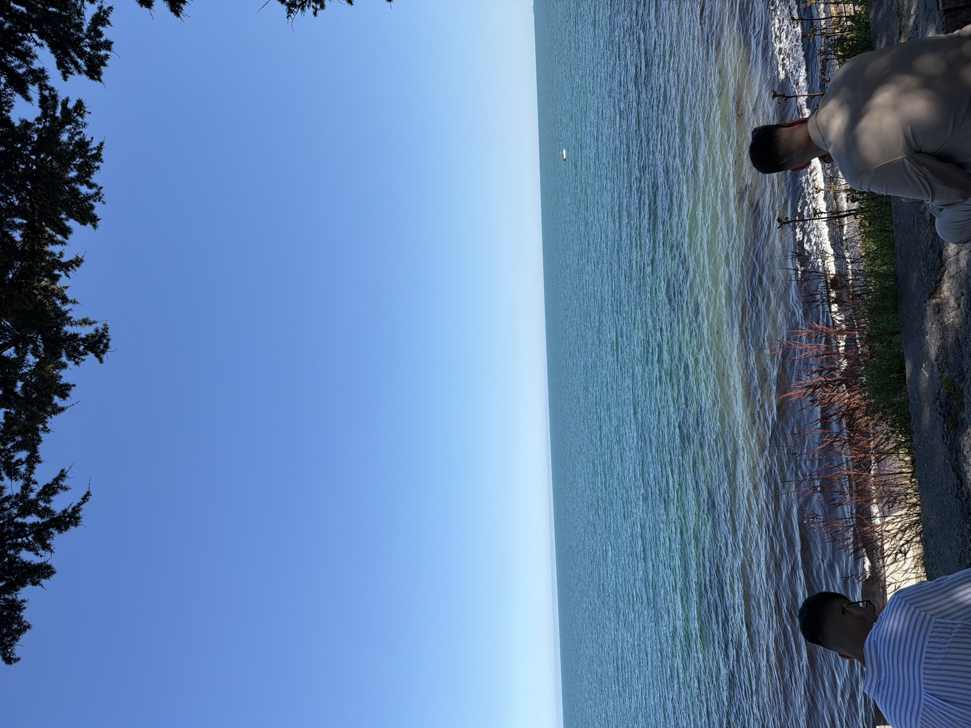 The image presents a vertically oriented shot with a scenic view. On the left, a large expanse of clear, blue sky dominates the frame, partially framed by the dark green foliage of trees in the upper left corner. The right side of the image features a body of water, likely a lake or sea, with visible surface waves and varying shades of blue and green. Along the edge of the water, there are plants and a paved walkway.
Two individuals are visible on the right side. One person is wearing a light-colored shirt and is kneeling near the edge of the water. Another person, wearing a blue and white striped shirt, is located at the bottom right corner, seemingly looking down at the water. The overall atmosphere is calm and serene. Given the mention of Nesebar, Bulgaria, it's reasonable to infer that this is a shoreline in that city, but without further cues I cannot confirm the specific location.