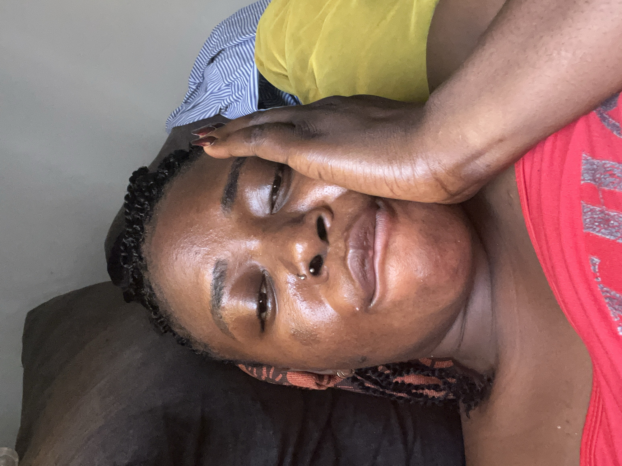 The media file features a close-up, portrait-style shot of a Black woman lying down. Her face is partially covered by her hand, which is resting on her forehead. She has dark braided hair, and a small nose ring is visible. Her eyes are partially closed, and her lips are slightly parted, suggesting a relaxed or contemplative mood.
She is wearing a red top with a patterned design and a blue and white striped garment is visible in the background, along with a yellow garment. A dark brown surface, possibly a pillow or mattress, is beneath her. The lighting appears to be natural and soft, suggesting it might be daytime. The overall scene conveys an intimate and personal moment. There is no visible text in the image. The location context is Abuja, Nigeria.
