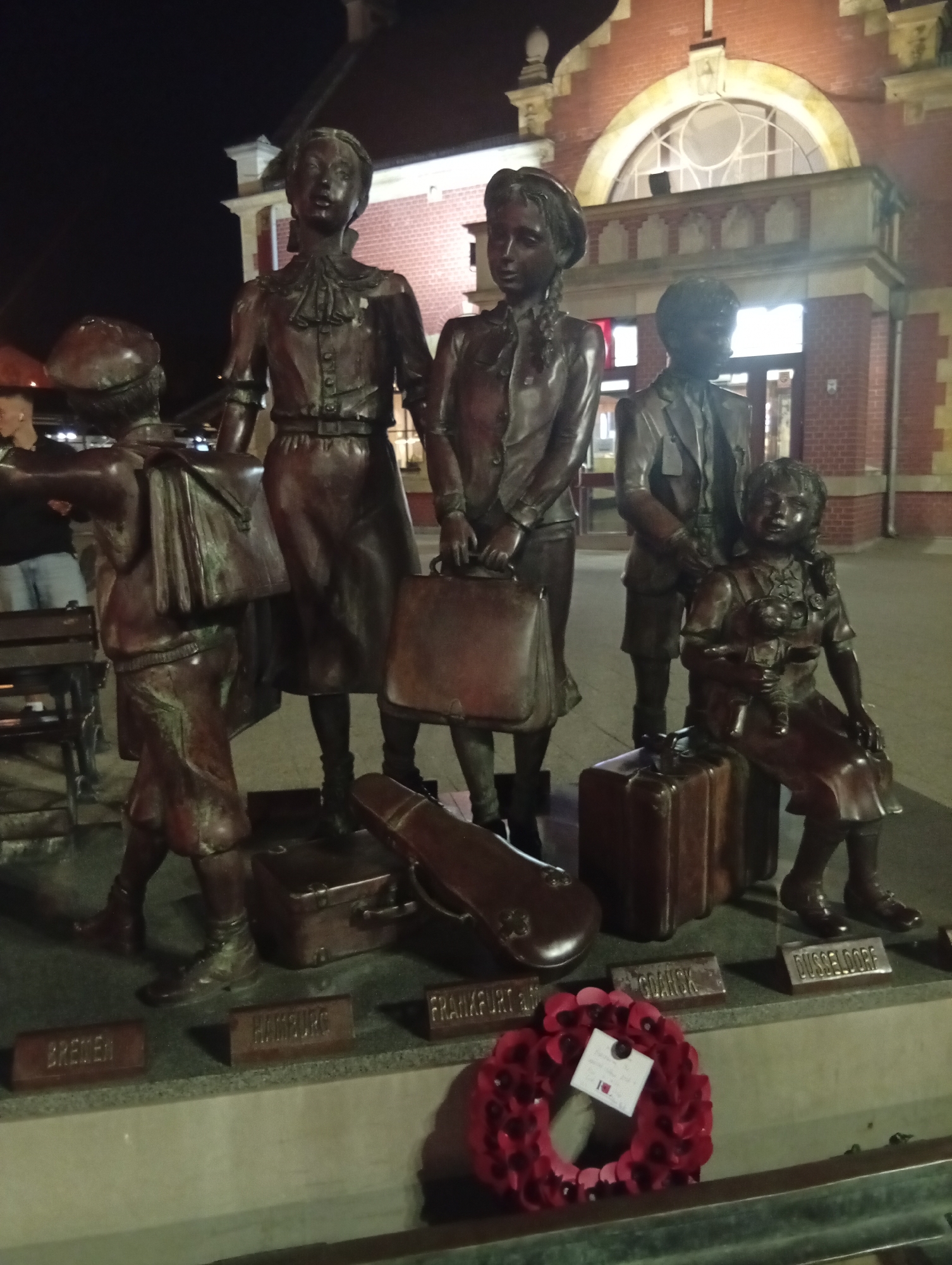 The image shows a night-time view of a bronze sculpture in Gdańsk, Poland, depicting children and their luggage. The sculpture, located near the Gdańsk Główny train station, commemorates the children who were evacuated from various German cities to safety during World War II.
In the foreground, a wreath of red poppies with a small white card is placed on a ledge. The sculpture itself features five bronze figures of children, varying in age and pose. One child carries a satchel over their shoulder, another holds a suitcase, and a young girl sits on a suitcase clutching a doll. Other suitcases and a violin case are scattered around the base of the figures. Small rectangular plaques bearing city names, such as "BREMEN," "HAMBURG," "FRANKFURT," "GDAŃSK," and "DÜSSELDORF," are visible in front of the sculpture, indicating the origins of the children.
The backdrop reveals the facade of a historic brick building, likely the train station, with arched windows and illuminated interiors. The overall scene is dimly lit, with artificial lights casting shadows and highlighting the bronze figures. The environment suggests an outdoor public space, likely a plaza or entryway to the train station.