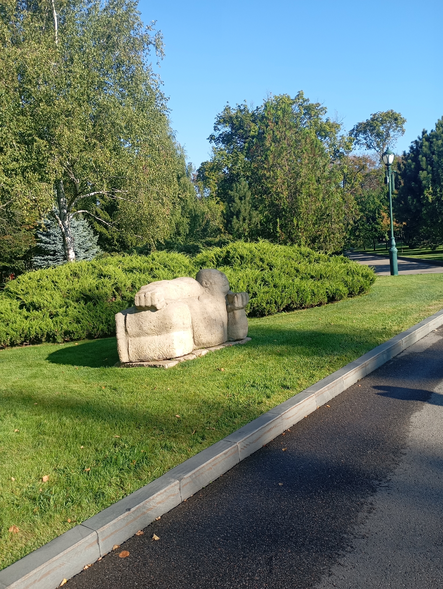 The image depicts a sunny outdoor scene in Kharkiv, Ukraine, likely in a park or garden. In the foreground, a paved walkway borders a well-maintained grassy area. A light-colored stone sculpture, abstract in form and appearing to represent a reclining figure, is positioned on the grass. Behind the sculpture, a dense, green hedge forms a backdrop. Further in the background, various trees, including evergreens and deciduous trees with green foliage, create a lush, natural setting. A street lamp with a distinctive design stands amongst the trees. The sky is clear and blue. The overall impression is one of a tranquil, scenic public space on a clear day.
