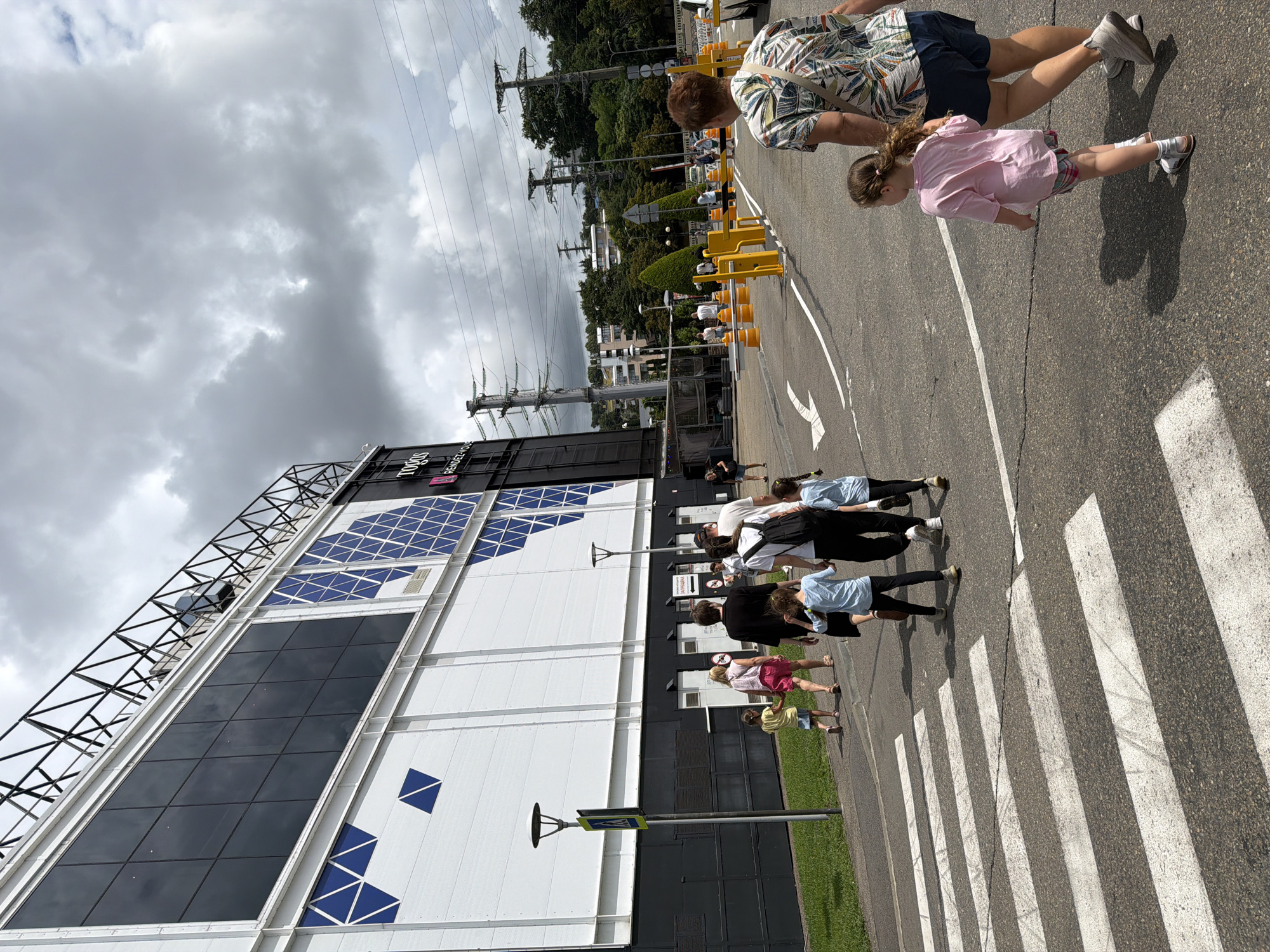 A group of people are walking on a paved area in front of a large modern building with a black and white facade and blue geometric patterns. The building has a prominent "TOD'S" logo. In the foreground, there are white crosswalk markings on the asphalt. Several individuals, including adults and children, are visible walking in various directions. One adult woman in a floral shirt and dark shorts is walking away from the camera with a young girl in a pink t-shirt and plaid shorts. Behind them, another group of people, including children, are walking towards the building. The environment appears to be an outdoor public space with trees and power lines visible in the background, under a cloudy sky. The location is identified as Sochi, Russia.