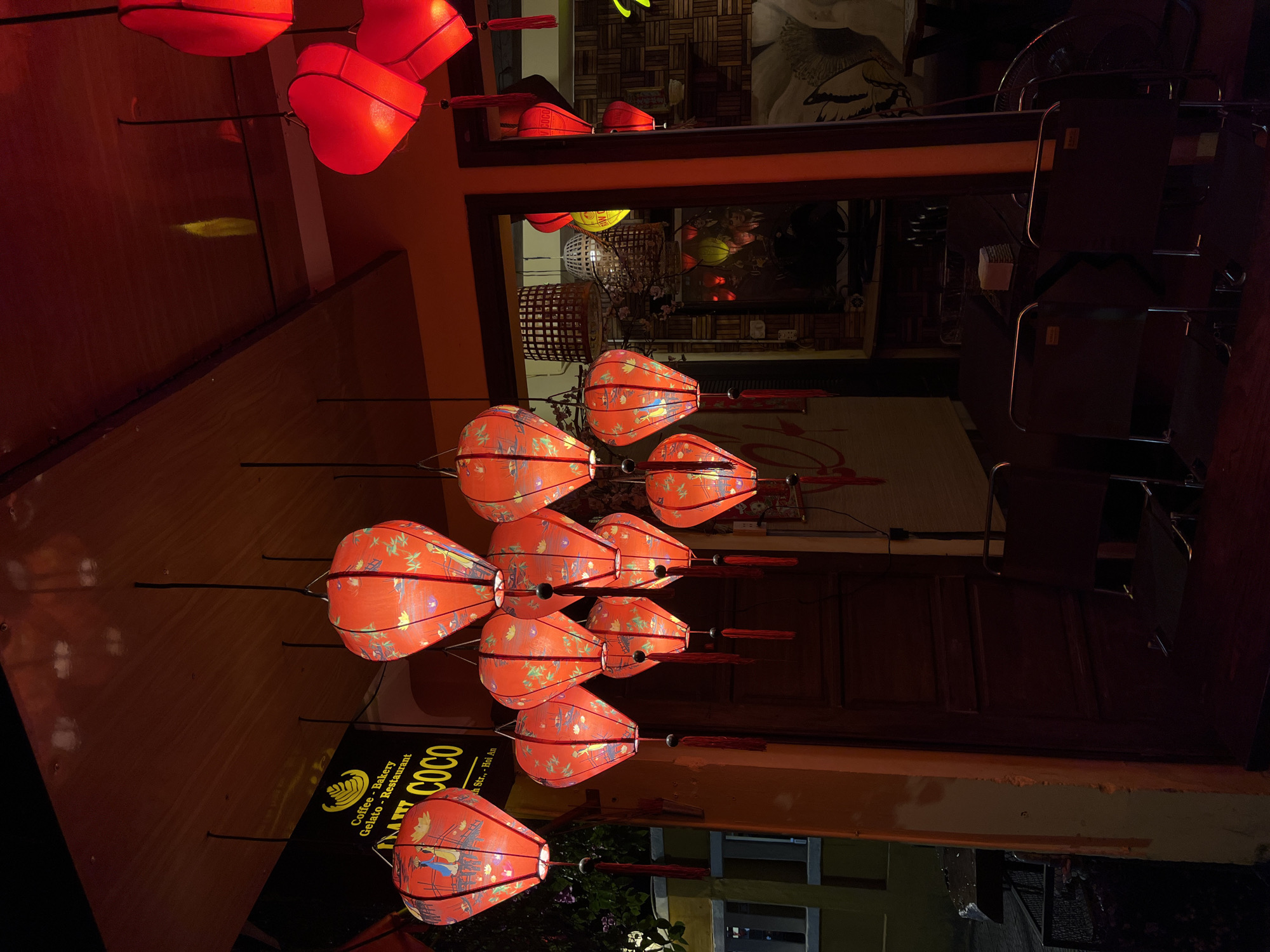 The image captures a street scene in Hội An Ward, Vietnam, featuring traditional red lanterns hanging in rows, their light illuminating the surrounding area. The lanterns are elongated and bulbous in shape, adorned with intricate patterns and designs.
A sign with the text "Coffee - Bakery Gelato - Restaurant THAT COCO" is visible, indicating a cafe or restaurant. The image also shows parts of buildings, including windows, walls, and a glimpse of greenery outside. The lighting is warm, adding to the ambience of the scene.