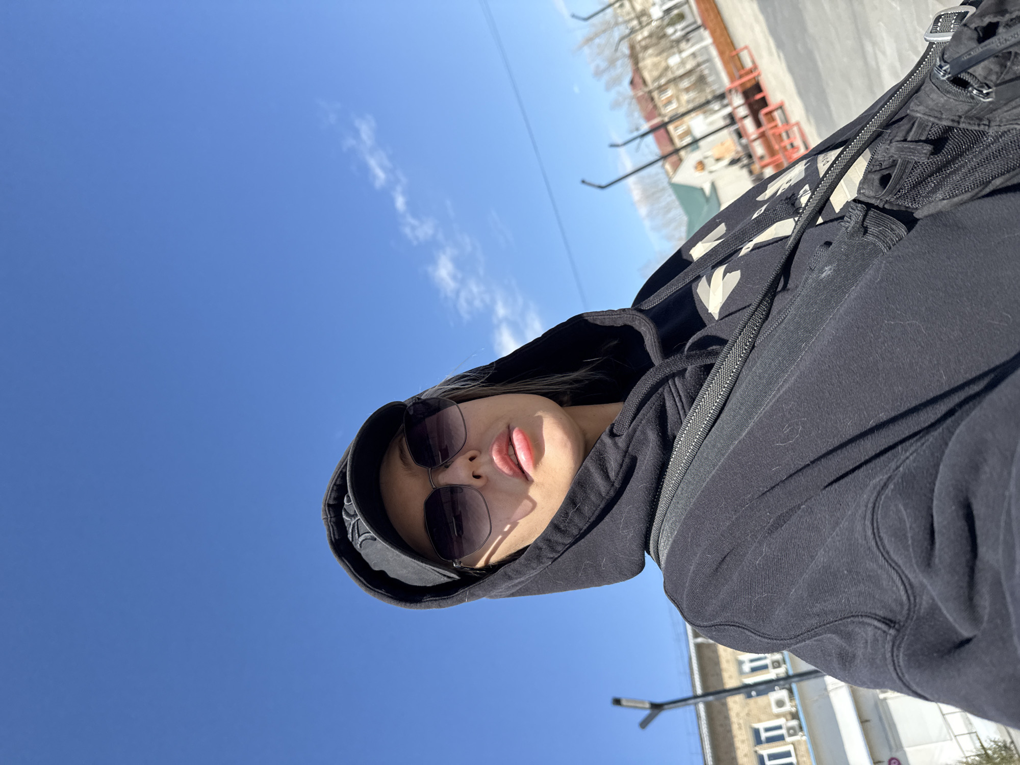 A low-angle, slightly canted, selfie of a woman with fair skin wearing a black hooded sweatshirt with white detailing and dark sunglasses. The background features a clear blue sky with a few scattered clouds above a cityscape. Buildings with multiple windows and structures resembling streetlights are visible in the distance. A portion of a backpack or bag with straps and buckles is visible. The overall scene suggests an outdoor environment. The geographical location is Borzya, Russia.
