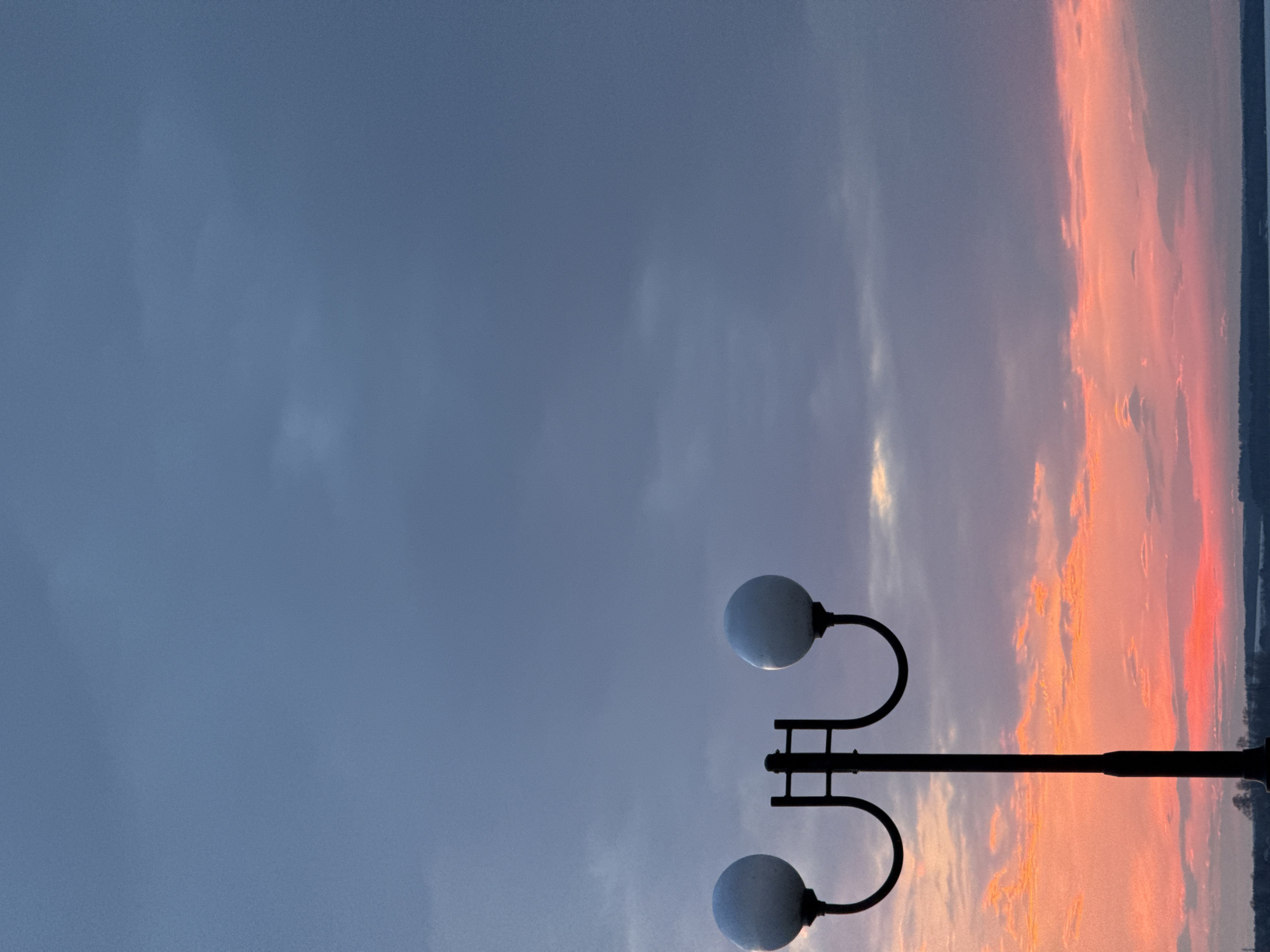 The image shows a dusk scene in Votkinsk, Russia. A street lamp with two spherical lights is silhouetted against a sky gradient ranging from dark blue-grey on the left to orange and pink hues of the sunset on the right. A line of dark land or water is visible at the far right edge of the image.