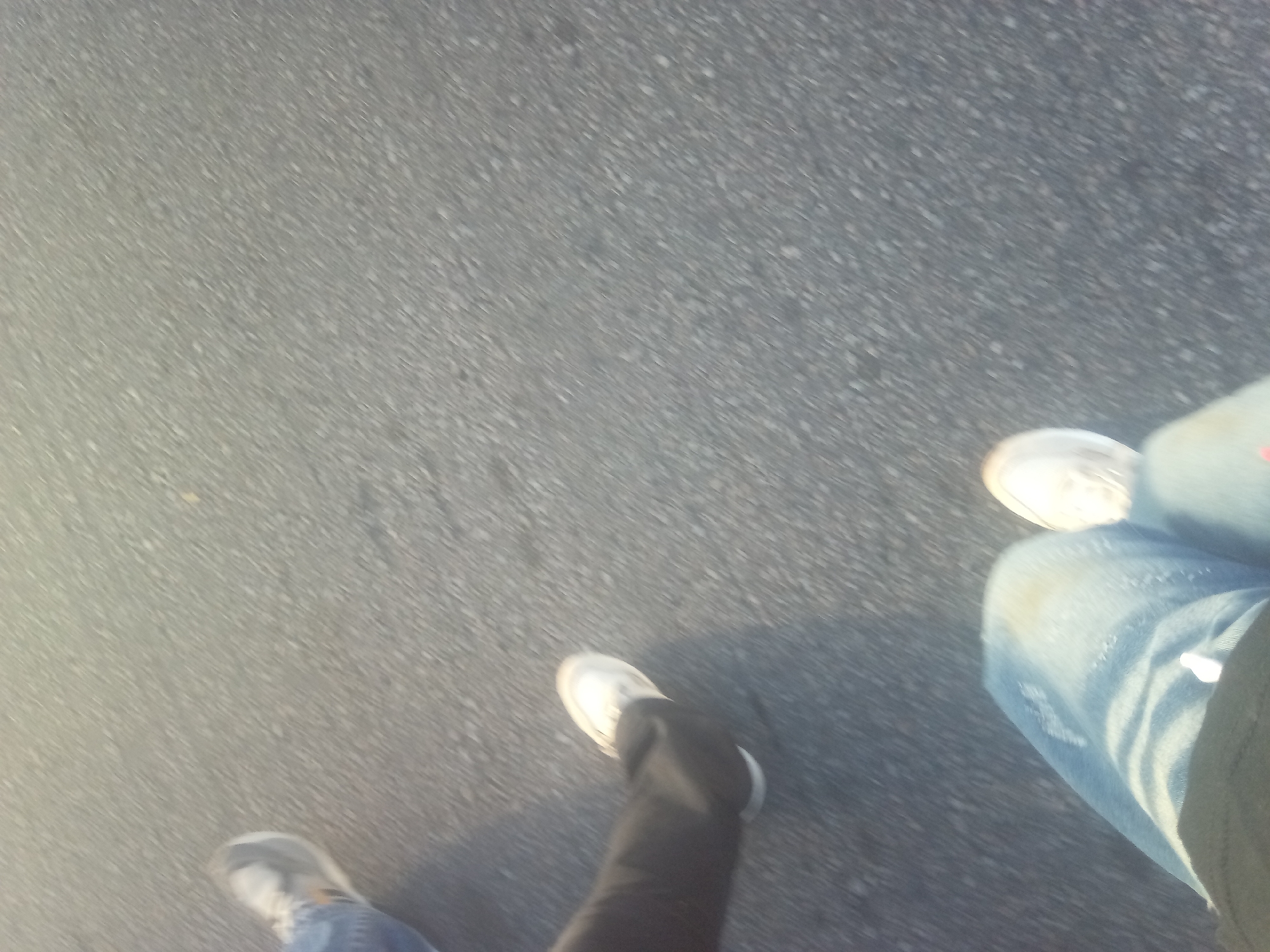 The image shows two people walking on asphalt. One person is wearing light blue jeans and white shoes, and the other is wearing dark pants and gray shoes. The asphalt appears to be a street or sidewalk. The location is not discernible from the image.