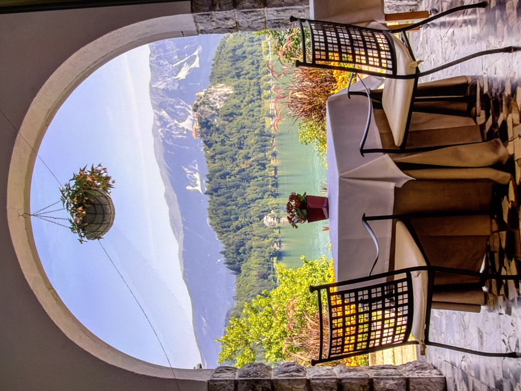 The image depicts a picturesque view of Lake Bled and Bled Castle in Slovenia, framed by an arched stone balcony. In the foreground, a table is set with a white tablecloth and two chairs with cream-colored cushions. A small pot of red flowers sits on the table. Beyond the balcony, a serene lake is visible, surrounded by lush green trees. In the distance, Bled Castle stands atop a rocky outcrop, with majestic mountains forming the backdrop. The sky is a clear blue. The scene is bathed in sunlight, suggesting a pleasant day. The architecture suggests a restaurant or cafe setting, offering diners a scenic vista.