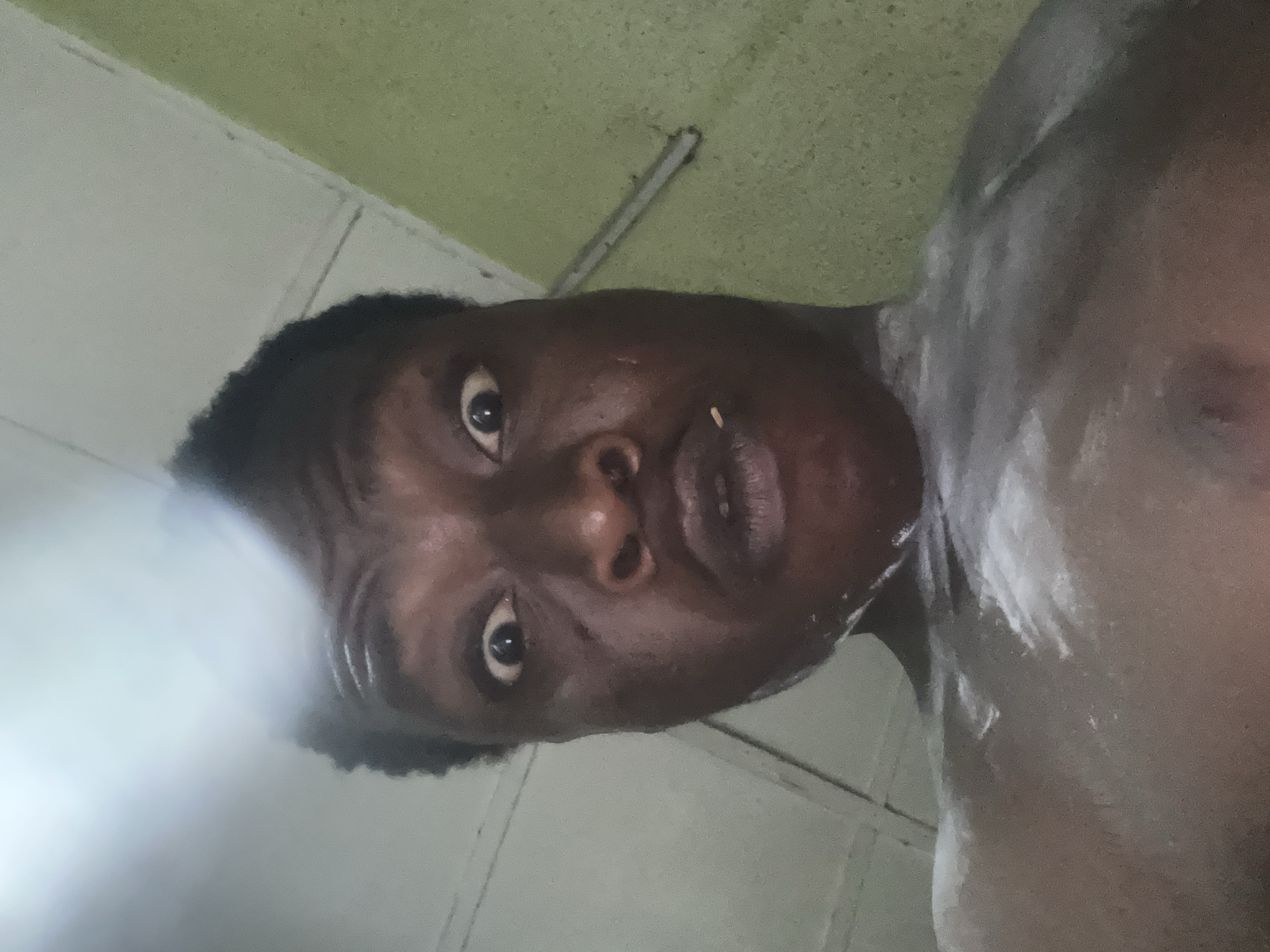 A close-up, low-angle portrait depicts a person with dark skin and short, dark, curly hair looking directly into the camera. Their eyes are wide and alert, and their mouth is slightly open, revealing a small, light-colored object on their lower lip or chin. A white, foamy or pasty substance is visible on the person's right shoulder and chest, suggesting they might be in the midst of washing or showering. They appear to be shirtless.
The background is an indoor setting with a light green wall or ceiling and some white panels or beams. The entire image is tilted, giving a disorienting perspective. A strong, hazy light source, possibly a camera flash, illuminates the lower-left portion of the frame, creating a bright glare. The rest of the scene is dimly lit. No text is visible in the image.