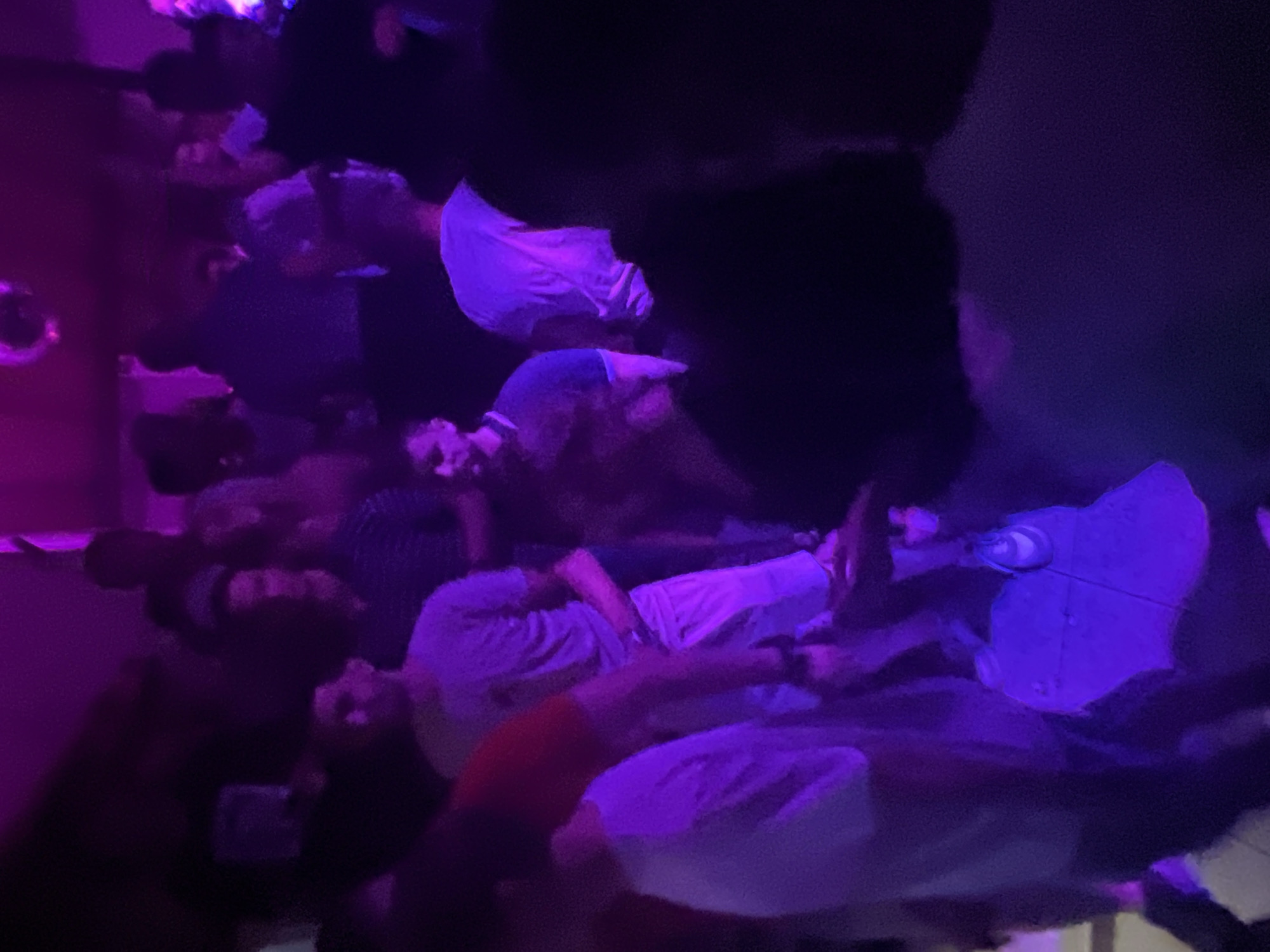 The image depicts a dimly lit nightclub scene with purple lighting. Several individuals are visible, with some appearing to be dancing or socializing. One person in the foreground is wearing a white t-shirt and shorts. Another individual is wearing a t-shirt with a dark collar. In the background, more people are gathered, with some silhouetted against the purple light. A disco ball is visible hanging from the ceiling. The scene appears to be an indoor venue, typical of a nightclub or bar. The specific location in Herceg Novi, Montenegro, cannot be determined from the visual information provided in the image.