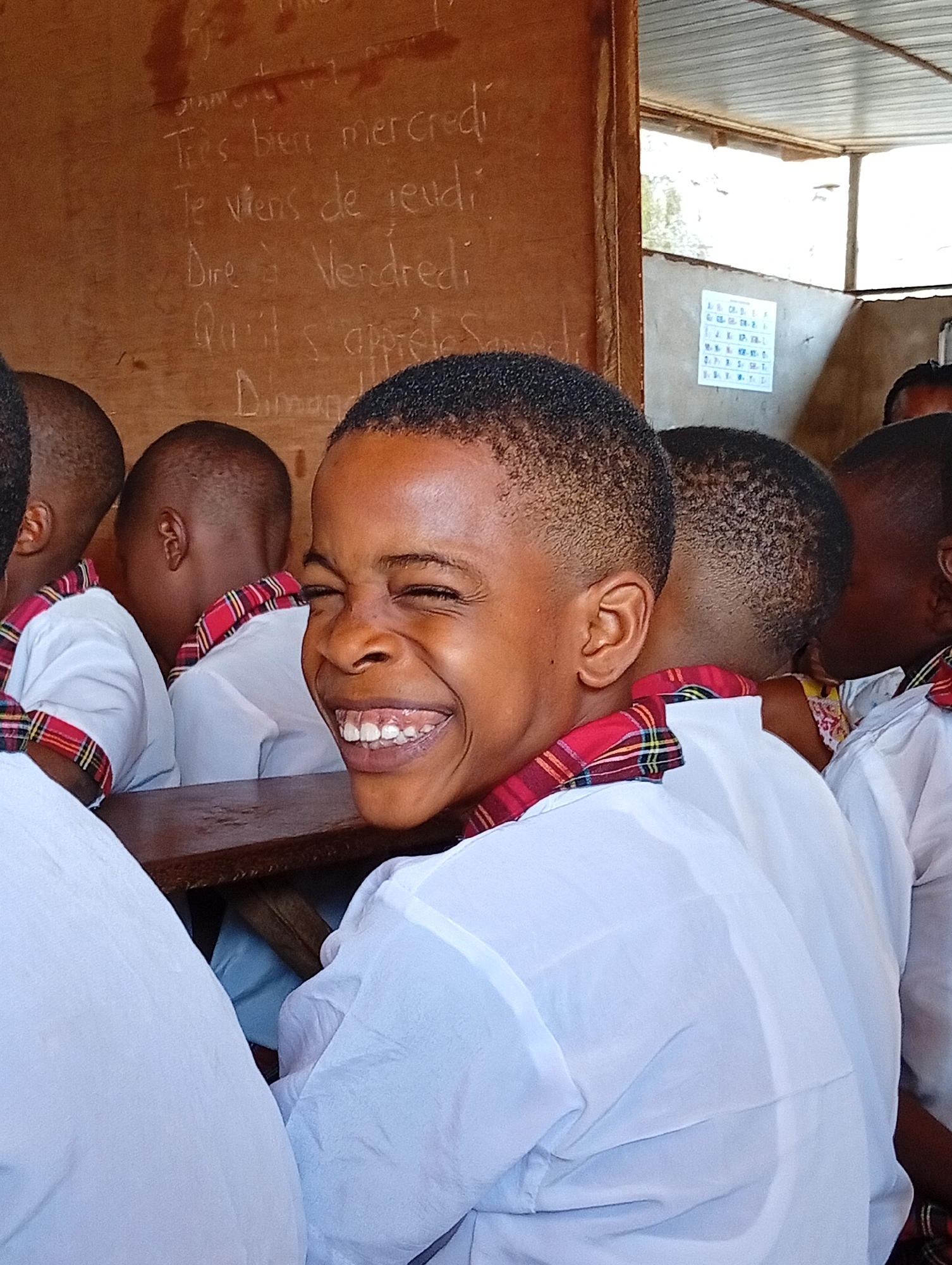 A young Black child in a white school uniform with a red and green plaid collar sits at a wooden desk, smiling widely and looking toward the camera. The child is in a classroom setting with other children in similar uniforms. A chalkboard with French text is visible behind them, along with a calendar. The picture appears to be taken indoors, possibly in a school in Ukwu-Abwa, Nigeria.