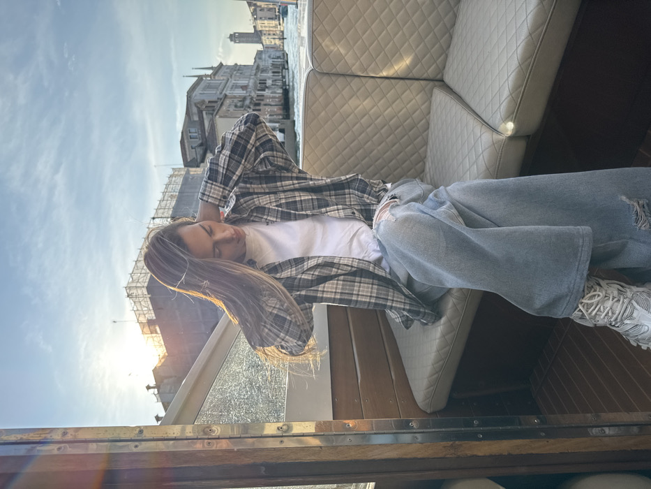 An individual with long, light-brown hair, wearing a white t-shirt, an open grey and white plaid shirt, light blue ripped wide-leg jeans, and white athletic shoes, is seated on a light-colored, diamond-quilted upholstered bench within a boat. The individual's head is tilted downwards, and both hands are positioned near the back of the head/neck area. The boat's interior features wooden paneling and a transparent window on the left. The background reveals a canal in Venice, Italy, flanked by historic multi-story Venetian buildings, one of which has visible scaffolding. A distant bell tower is visible on the right horizon. The sky is partly cloudy, and a low sun positioned on the left side of the frame creates a bright glare and lens flare.