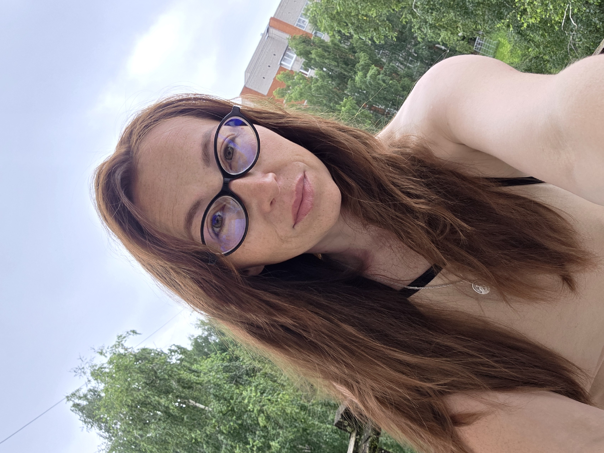 A woman with long, auburn hair and glasses is taking a selfie outdoors. She is wearing a beige top and a silver pendant. Behind her, trees and a building are visible. The image appears to have been taken in Chaikovsky, Russia.