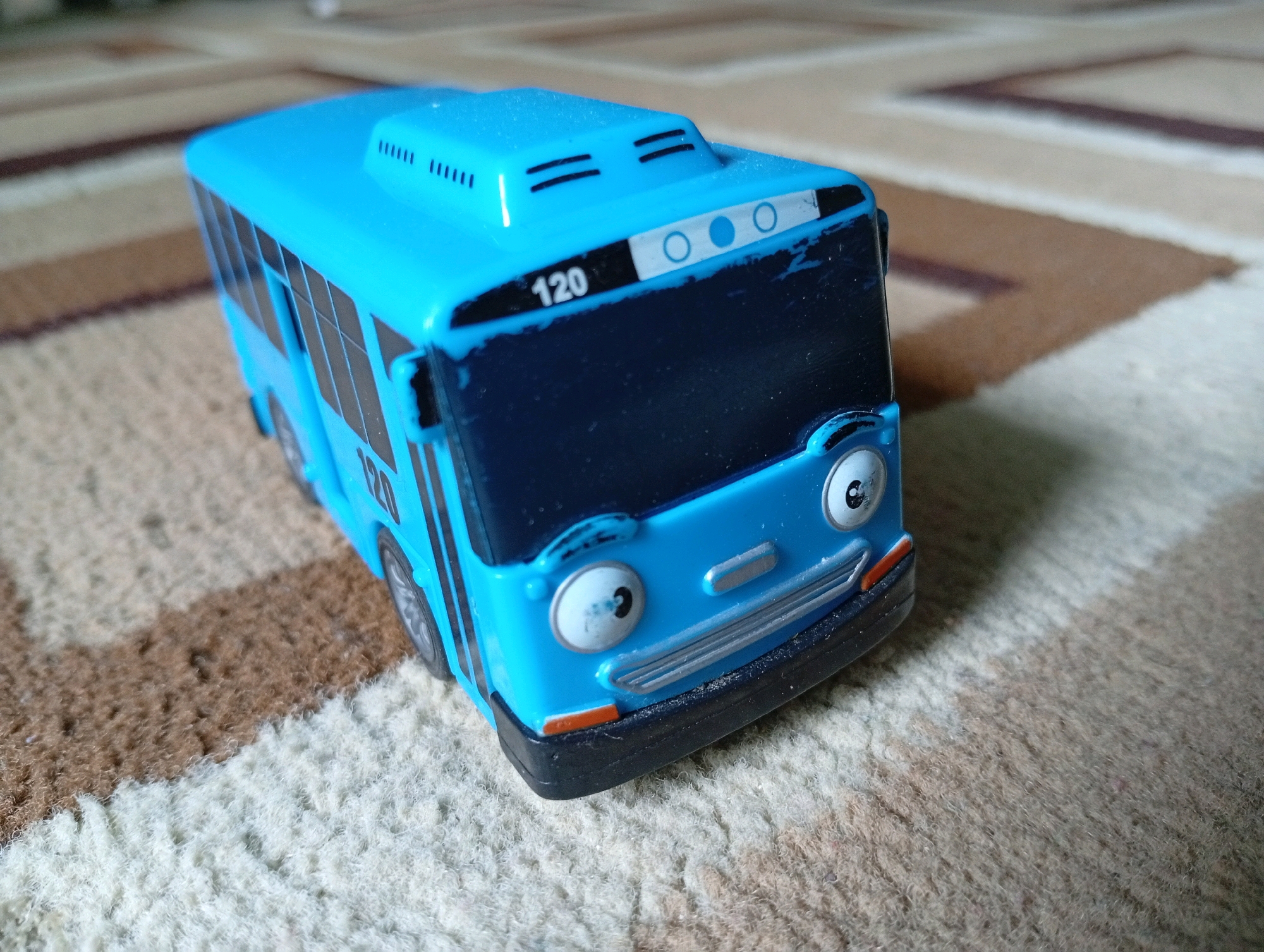 A close-up, eye-level shot shows a bright blue toy bus with a friendly face, sitting on a carpet. The bus is angled towards the viewer, with its front end slightly off-center to the right. The carpet features a striped pattern of light beige, brown, and white. The bus has black windows, two large round eyes with black pupils, and a smiling mouth. A silver grille and orange turn signals are visible on the front. The number "120" is displayed prominently on the front of the bus, near the roof. The roof of the bus has several black horizontal lines. The scene is indoors, and no people are visible. The overall impression is one of a clean, well-lit indoor environment.