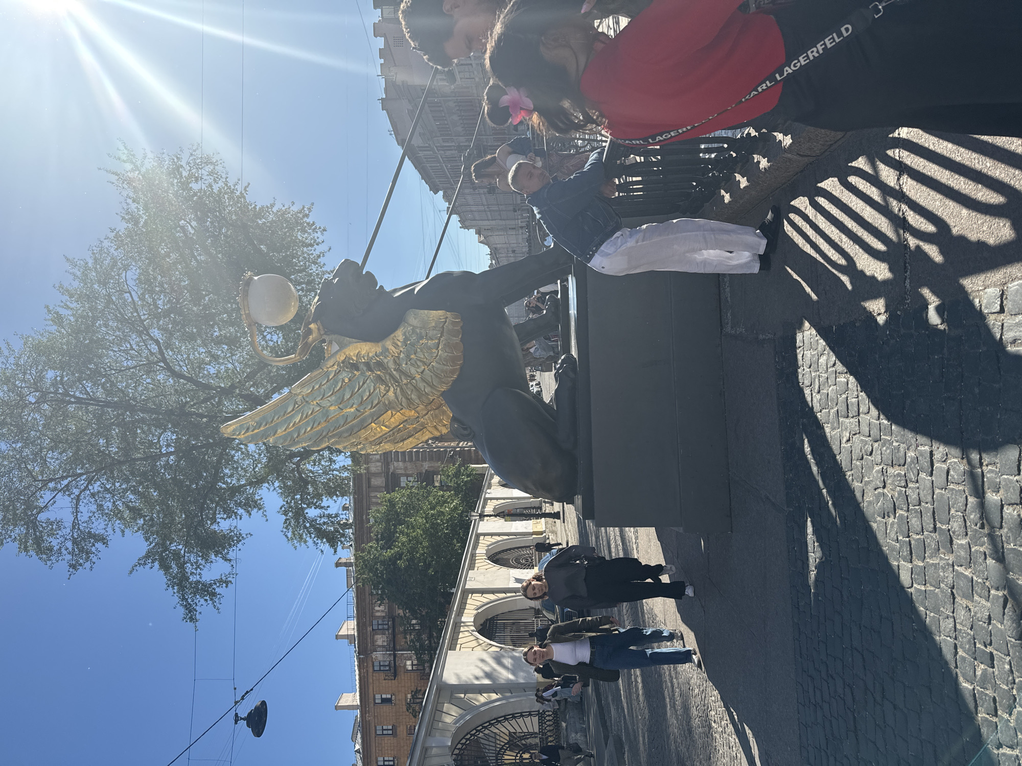 The image captures a sunny day in Saint Petersburg, Russia, with a bright sun casting long shadows across a cobblestone street. In the foreground, a bronze statue of a winged creature, possibly a sphinx or griffin, stands prominently. The creature is depicted with golden wings and holds a round object, perhaps a globe or orb.
Several people are visible, some standing near the statue and others walking along the street. They appear to be tourists or locals enjoying the weather. One woman in the background wears a red top and black pants with "KARL LAGERFELD" written on the waistband. The people seem relaxed, possibly engaged in sightseeing or casual conversation.
The background reveals the architecture of Saint Petersburg, with buildings featuring classical elements like arched windows and ornate detailing. A large tree with green foliage is also visible, contributing to the pleasant outdoor atmosphere. The bright sunlight and clear blue sky suggest it is daytime, likely mid-morning or early afternoon. The overall scene conveys a sense of everyday life and tourism in a historic European city.