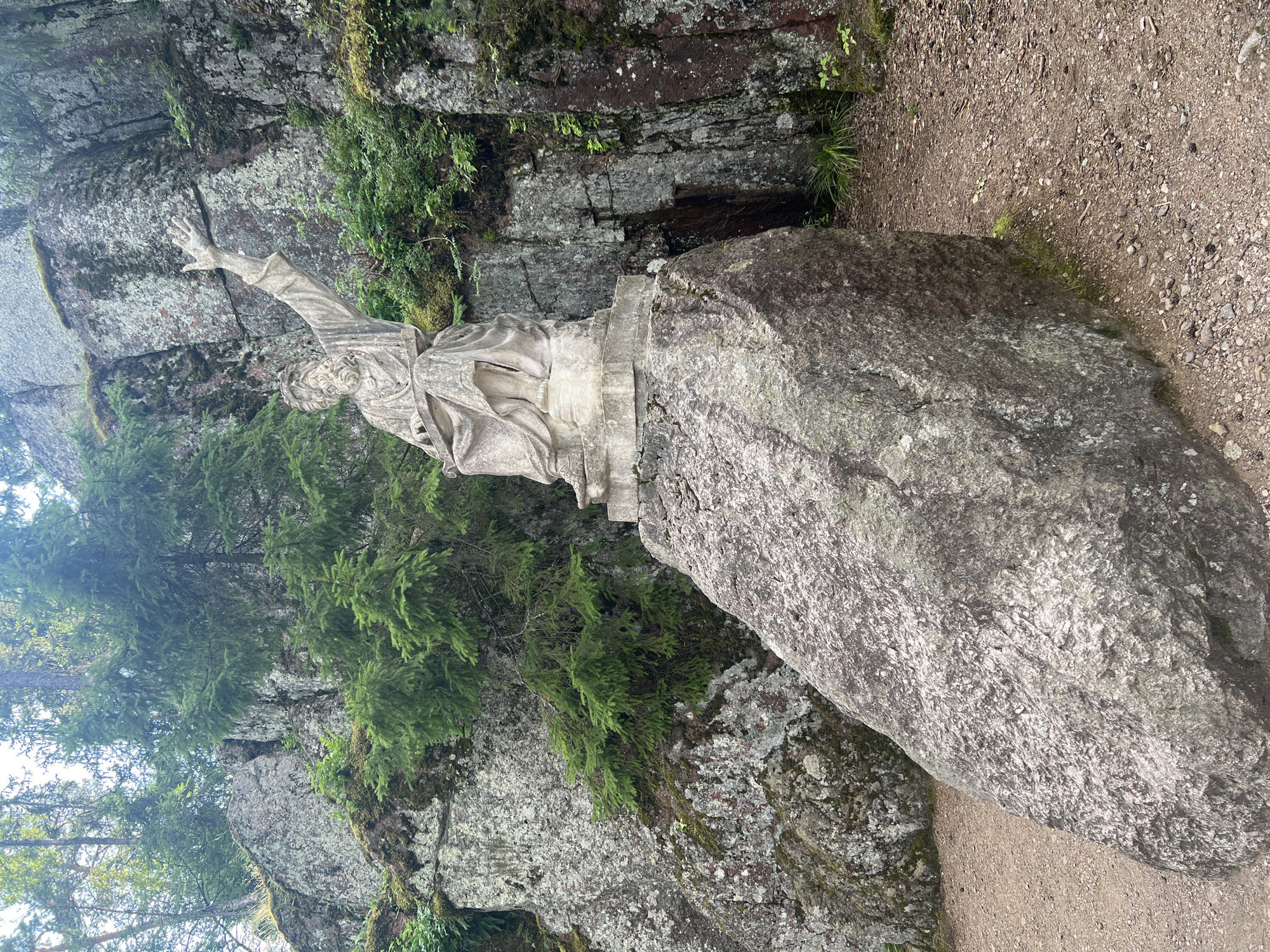 A stone statue of a man with a beard and flowing robes stands on a large rock base. He is seated, with his right arm raised and extended, fingers spread. His left hand rests on a book or tablet in his lap. The statue is situated in a rocky, wooded area with trees and moss-covered rocks visible. The ground around the statue is a dirt path scattered with small stones and pine needles. The scene appears to be outdoors in a natural setting. This statue is located in the Mon Repos Park in Vyborg, Russia.