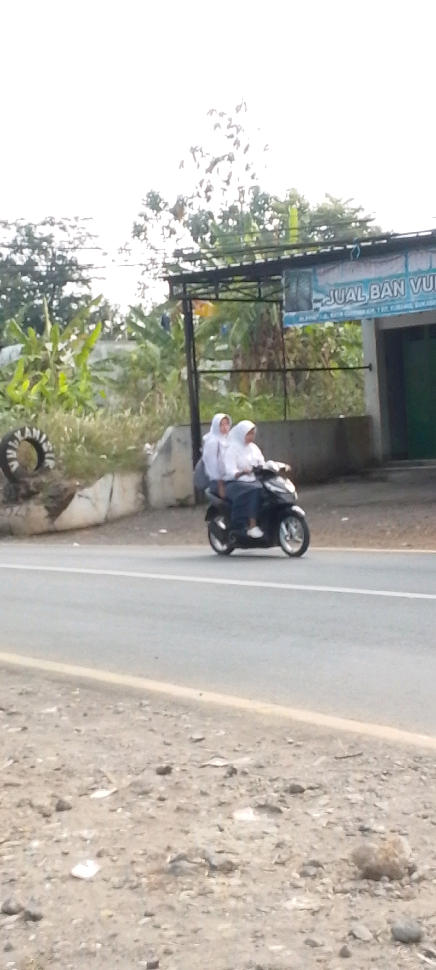 Two young individuals in white headscarves and white shirts are riding on a black motorcycle. The individual in front is operating the motorcycle, while the individual behind them is a passenger. They are traveling on a paved road in an outdoor environment, with a slightly hazy sky. To the left of the road, there is a tire with writing on it and a white wall. Further in the background, a building with a blue banner displaying text and some green doors is visible, along with lush green foliage and trees. The scene appears to be in the city of Cilaku, Indonesia, based on contextual information.