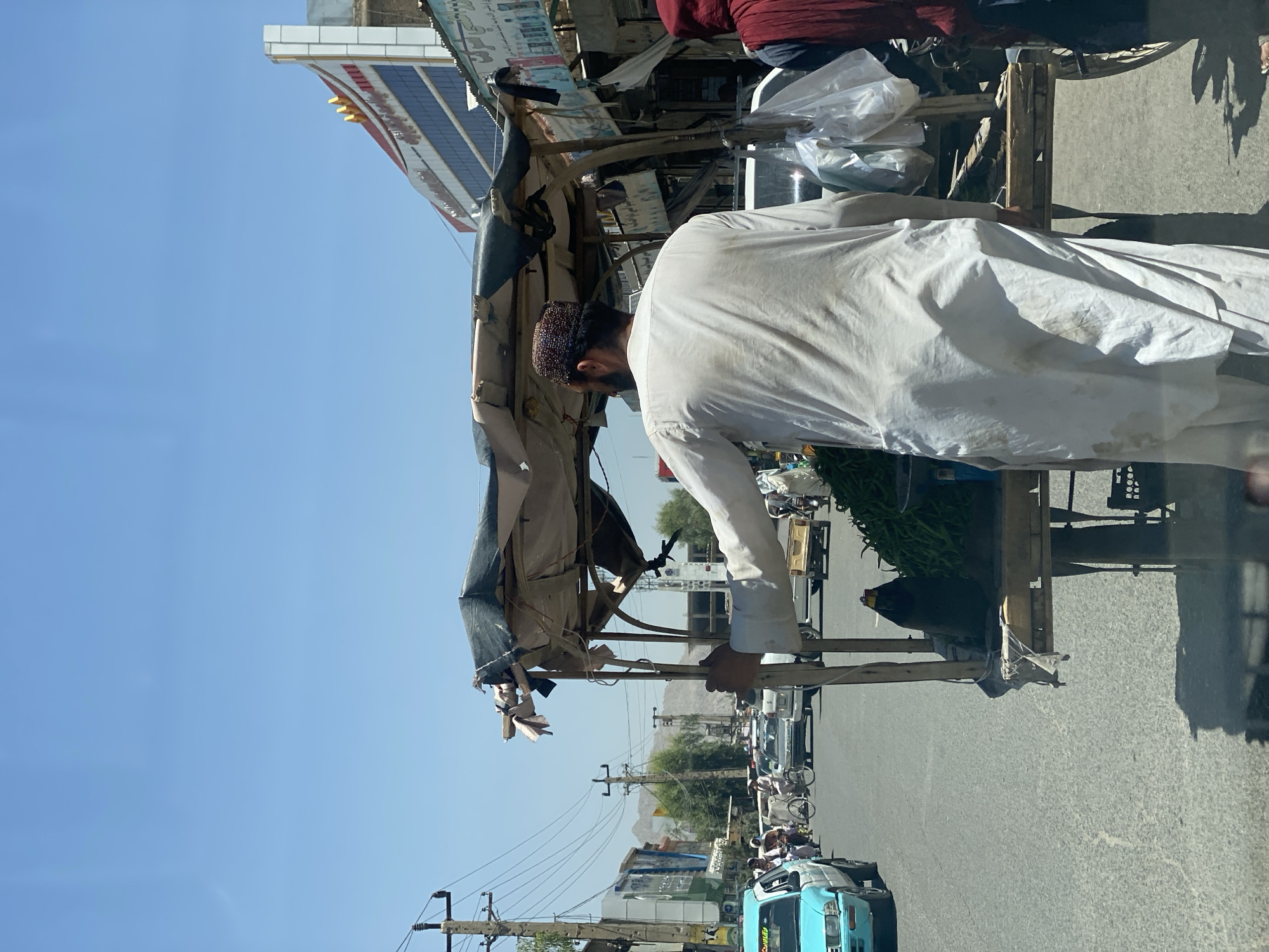 In Kandahar, Afghanistan, a street scene unfolds under a clear blue sky. A man, dressed in a white tunic and a patterned cap, stands behind a cart laden with green vegetables, possibly chilies, and two dark bottles. He appears to be a vendor, his hand resting on the cart's frame as he looks to his left. Beside him, another individual in a red head covering stands partially visible. The cart, partially covered by a worn, patched awning, is positioned on a paved street filled with traffic. A light blue vehicle is prominent on the left, alongside other cars and a bicycle. In the background, buildings line the street, one featuring a prominent sign with red and gold lettering, hinting at commercial activity. Power lines crisscross the sky, and distant mountains are visible, suggesting the city's geographical setting. The overall atmosphere is one of a bustling marketplace in a sun-drenched urban environment.