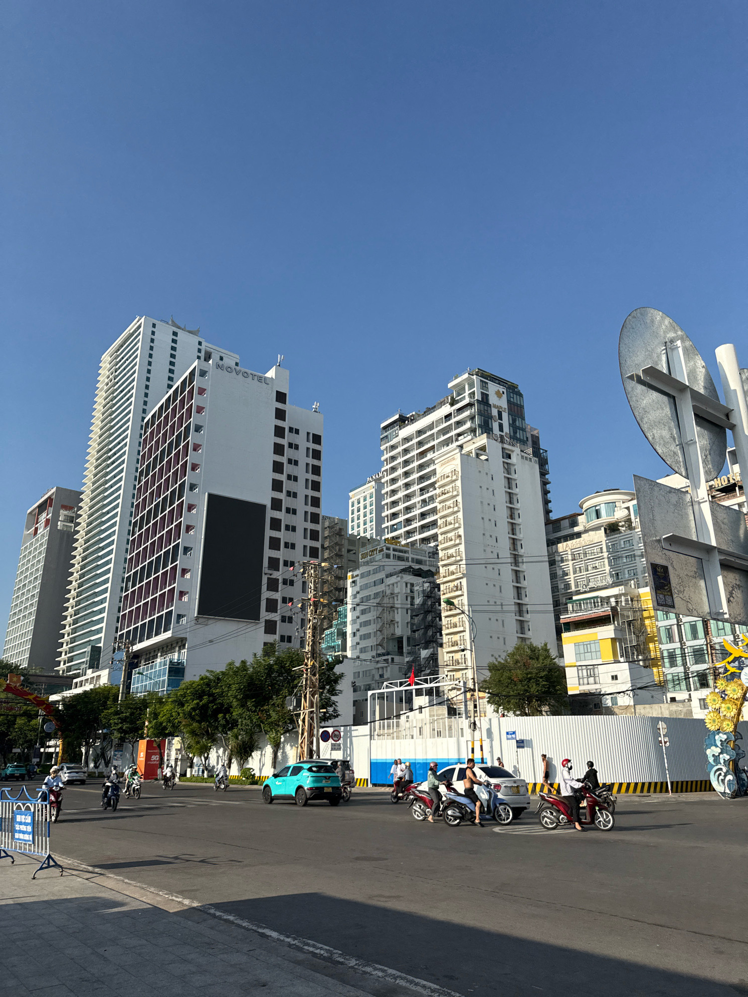 This is a wide-angle, street-level photograph taken in Nha Trang, Vietnam, capturing a busy intersection with numerous buildings and ongoing traffic. The sky is a clear, bright blue.
Dominating the background are several tall, modern buildings, some featuring glass facades and balconies. One prominent white building displays the "NOVOTEL" logo. Another building has a large, dark rectangular screen on its facade. The architecture suggests a commercial or hotel district.
In the foreground, the paved street is filled with motorbikes, a common mode of transportation in Vietnam. Several individuals are riding these motorbikes, some wearing helmets. A turquoise car is also visible on the road. The traffic appears to be moving, indicating normal activity.
On the sidewalk, there are some trees and decorative elements, including a yellow and gold ornamental structure with floral motifs on the right side of the frame, possibly for a festive occasion. Temporary blue and white barriers are present on the left, suggesting a construction or event area. Street signs and utility poles with visible wiring are also part of the urban landscape.
The lighting suggests it is daytime, with clear shadows cast on the road, indicating the sun is high. The overall impression is of a vibrant, active city scene.