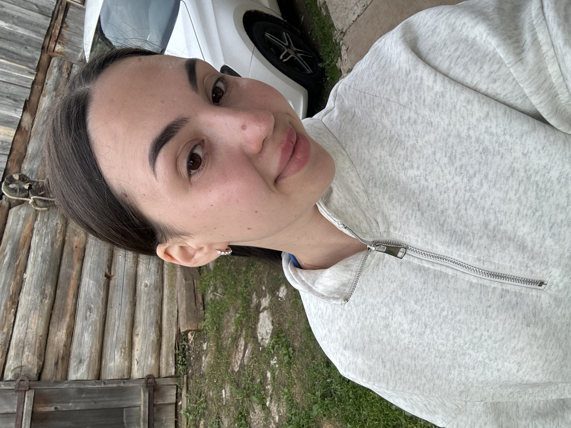 A woman is photographed in front of a wooden structure and grass. A partial view of a white car with a black tire is visible in the background. The woman is wearing a light gray zip-up jacket. She appears to be smiling slightly and looking at the camera. The location cannot be determined from this image.
