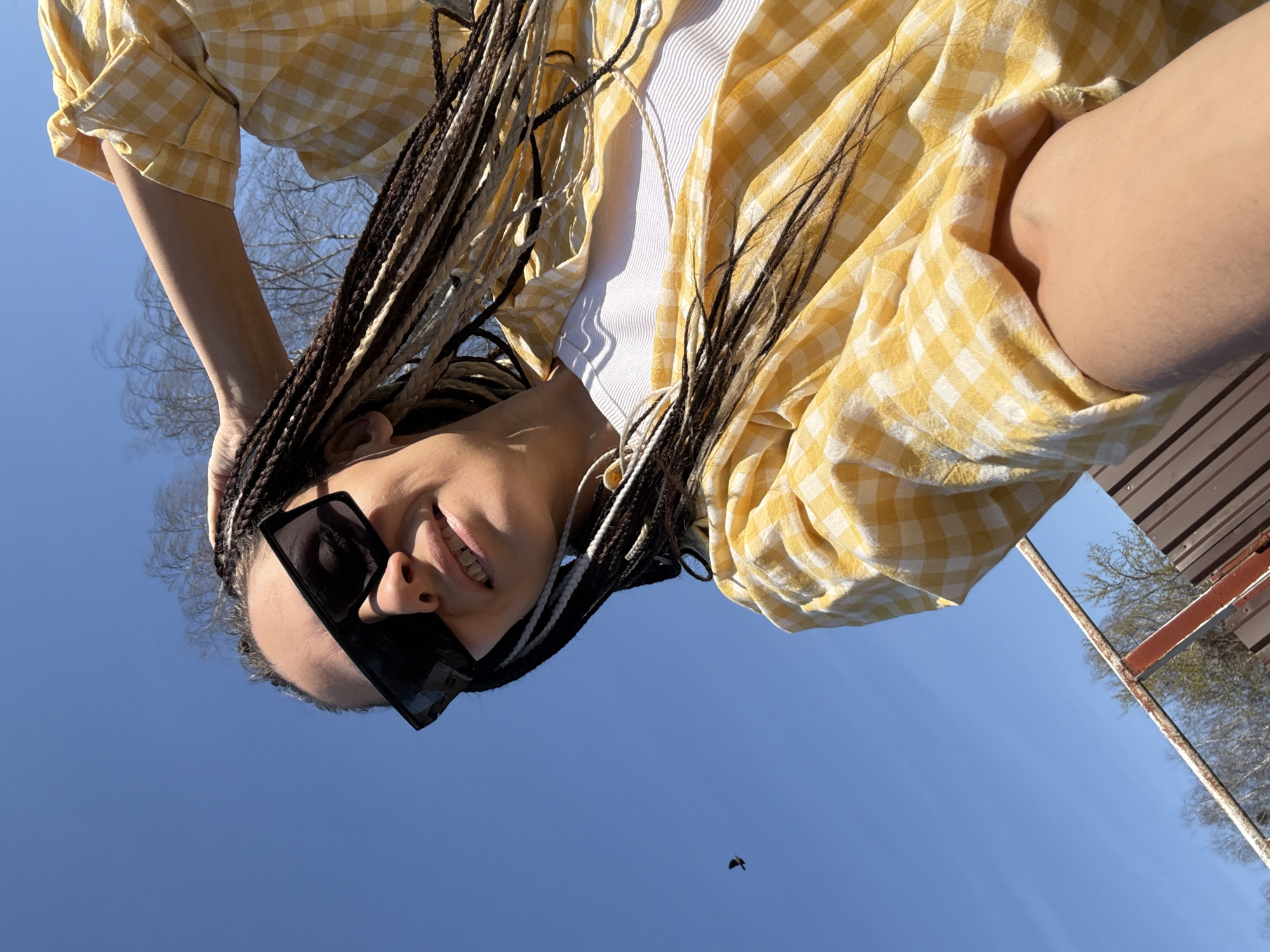 A woman with braided hair and dark sunglasses looks down at the camera. She is wearing a white top and a yellow and white checkered shirt. She is outdoors, with a clear blue sky visible in the background. A bird is flying in the lower center of the frame. The setting appears to be Fedorino, Russia, based on the image's origin.