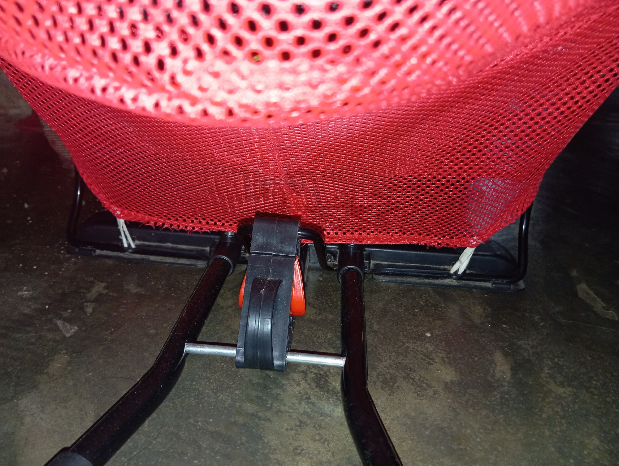 A close-up, low-angle shot shows the base of a red mesh basket attached to a black metal frame. The basket is made of a coarse, red mesh with visible holes, and is supported by a frame with two angled legs. A black plastic mechanism with an orange lever is centered on the frame, connecting the legs to the basket's support. White ties are visible securing the basket to the frame. The scene appears to be indoors, on a dark, textured floor that may be concrete, with some scattered dust. The lighting is dim, suggesting an indoor environment possibly at night. There are no people or discernible actions in the image. The location is specified as Rejosari, Indonesia, but there are no visual cues within the image to confirm this location.
