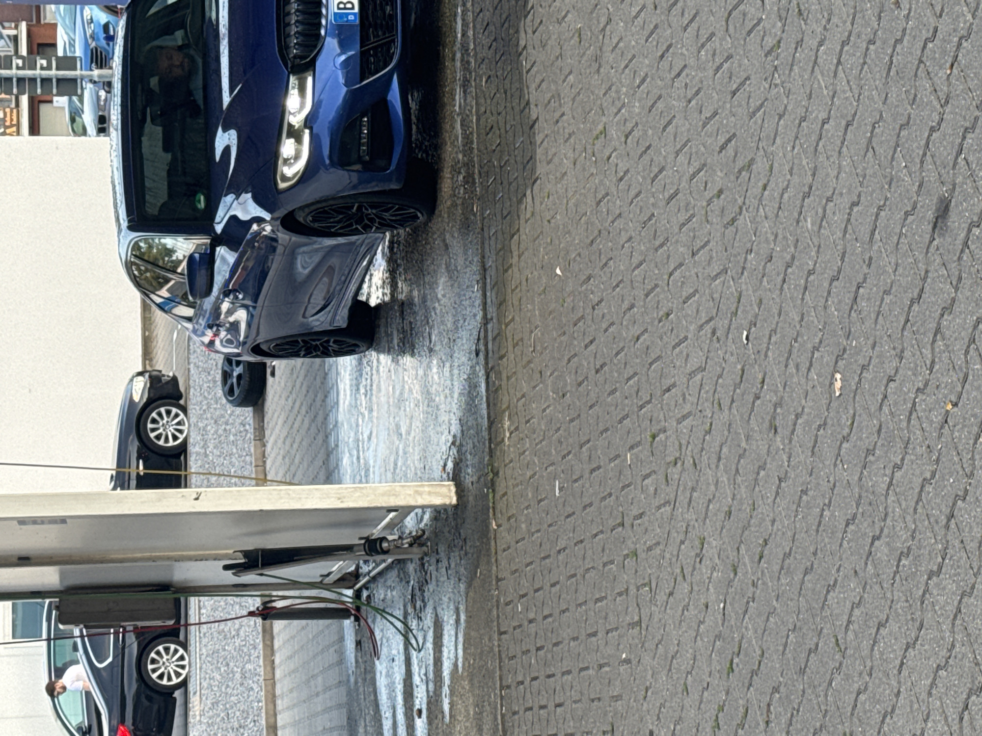 The image depicts a street scene, likely in Ahlen, Germany, featuring a blue BMW sedan being washed at a self-service car wash. A black BMW sedan is parked to the left, with a person wearing a white shirt standing near it. The ground is paved with interlocking concrete blocks, and water from the car wash has created wet patches. In the background, other vehicles and buildings are visible, suggesting an urban setting. The blue BMW is the primary subject, showcasing its headlights and front grille. The presence of car wash equipment, like hoses and a water spraying apparatus, indicates the ongoing activity.