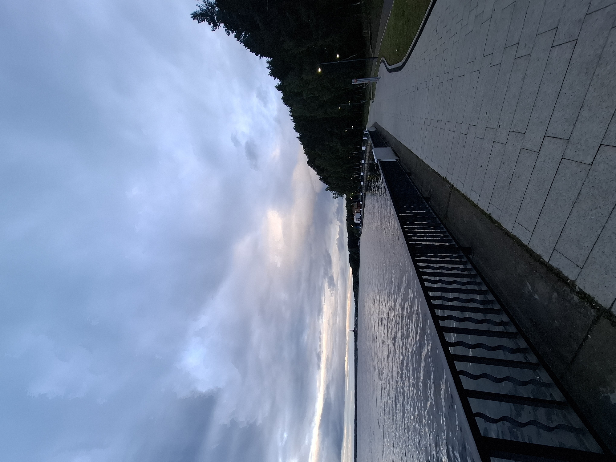 The image captures a tranquil evening scene along a waterfront promenade in Chaikovsky, Russia, likely bordering the Kama Reservoir. The sky is largely overcast with dense, stratified grey clouds, revealing a faint strip of lighter, desaturated orange and pink hues near the horizon, indicative of twilight. A vast body of dark, rippled water occupies the left half of the frame, extending towards the distant horizon. Across the water, two prominent, slender industrial chimneys or towers are visible against the cloudy sky.
In the foreground on the left, a dark metal railing with regularly spaced, undulating vertical bars runs parallel to the water's edge, anchored to a concrete base. Adjacent to the railing, a wide, light-grey paved walkway extends from the foreground into the midground, curving gently to the right. The walkway is constructed from rectangular and trapezoidal paver stones arranged in a staggered pattern. To the right of the walkway, a narrow strip of neatly maintained green grass is present.
A dense line of tall evergreen trees, predominantly pines, borders the right side of the walkway, forming a dark, imposing forest edge that recedes into the distance. Multiple modern streetlights, each with a luminous globe, are positioned at intervals along the tree line, casting light onto the path. Further along the walkway in the midground, some low-rise structures with light-colored walls and reddish roofs are discernible near the water. No individuals are visible on the promenade or in the water. The scene conveys a sense of quietude and twilight illumination.