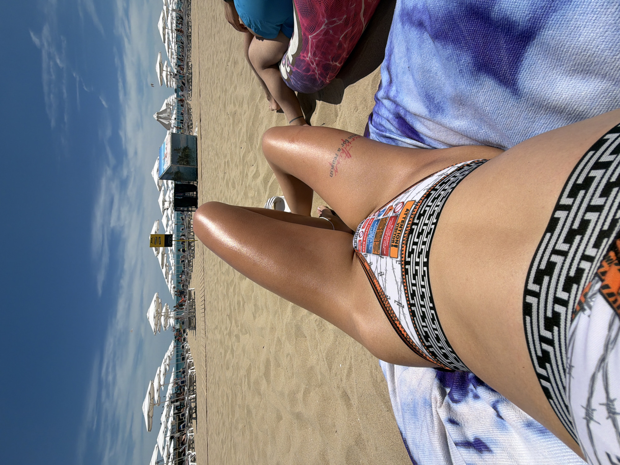 This is a first-person perspective shot of a person lying on a beach in Nesebar, Bulgaria. The person is wearing a bikini with a white background and black, orange, and white geometric patterns. A tattoo is visible on the upper thigh, appearing to say "This is for a Reason." The person is lying on a tie-dye towel with blue and purple hues. To the right, another person is visible, wearing blue shorts and lying on a pink and black inflatable. The background shows a sandy beach with rows of white lounge chairs and umbrellas. Further back, a wooden walkway is visible, as are small structures and a sign. The sky is blue with scattered white clouds. The sun is shining brightly, casting shadows.