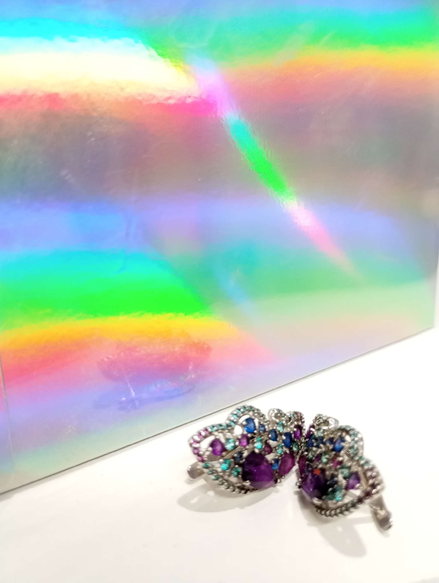 A close-up shot shows a pair of silver-colored earrings with a purple, tear-shaped gemstone at the center of each. The earrings are also adorned with smaller blue and teal gems around the central stone. The background is a reflective, holographic surface showing a gradient of colors, including blue, green, yellow, pink, and silver. The earrings are placed on a white surface, which is likely a table or countertop.
This image does not contain information that could directly link it to the city of Pervomaysky, Russia, or any specific location. It is a product shot of jewelry against a decorative background.