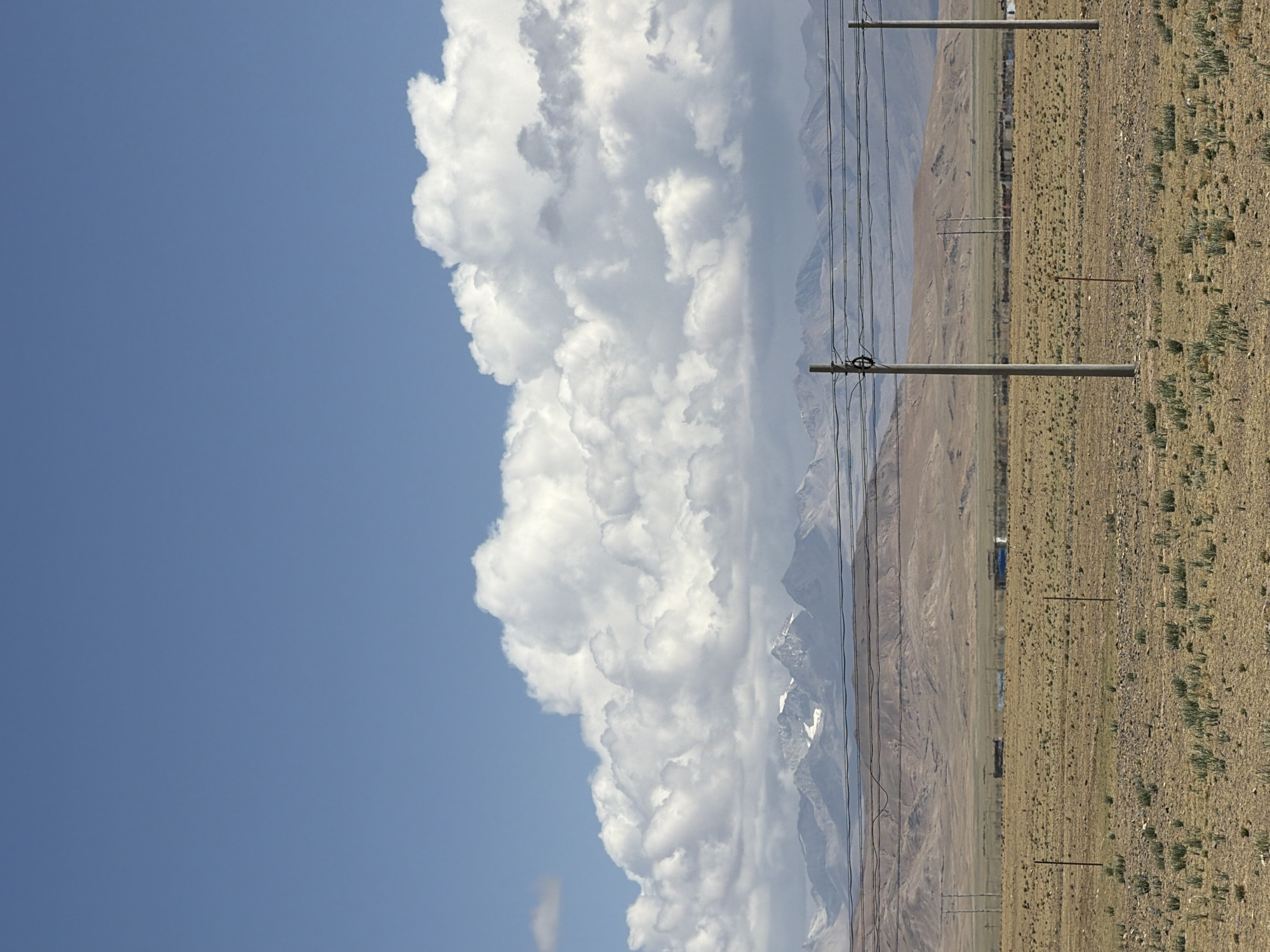 The image presents a vast, high-altitude landscape under a bright, sunny sky. The upper left portion of the frame features a clear, deep blue sky. Dominating the middle section are large, puffy white cumulus clouds, brilliantly lit by the sun, stretching across the sky and extending downwards towards the horizon.
Below the cloud formation and across the mid-ground, a rugged mountain range is visible. Some of the higher peaks are capped with snow or glaciers, suggesting significant elevation. The mountains display earthy, rocky textures.
In the foreground and extending into the mid-distance is an expansive, dry, and undulating plain. This land is covered with sparse, low-lying yellowish-brown vegetation, characteristic of an arid or semi-arid environment. Several utility poles, likely for power or communication, are planted across this plain, with numerous dark wires strung between them, running generally parallel to the mountain range. One prominent pole in the foreground clearly shows its concrete-like structure and multiple wires. In the far distance, near the base of the mountains, a few indistinct structures or buildings can be faintly discerned.
The scene depicts bright daylight, likely midday, given the strong illumination of the clouds and landscape. The weather appears clear and sunny, with distinct cloud formations. The overall impression is one of natural grandeur and remoteness, with human infrastructure integrated into the vast environment. There is no visible text in the image.