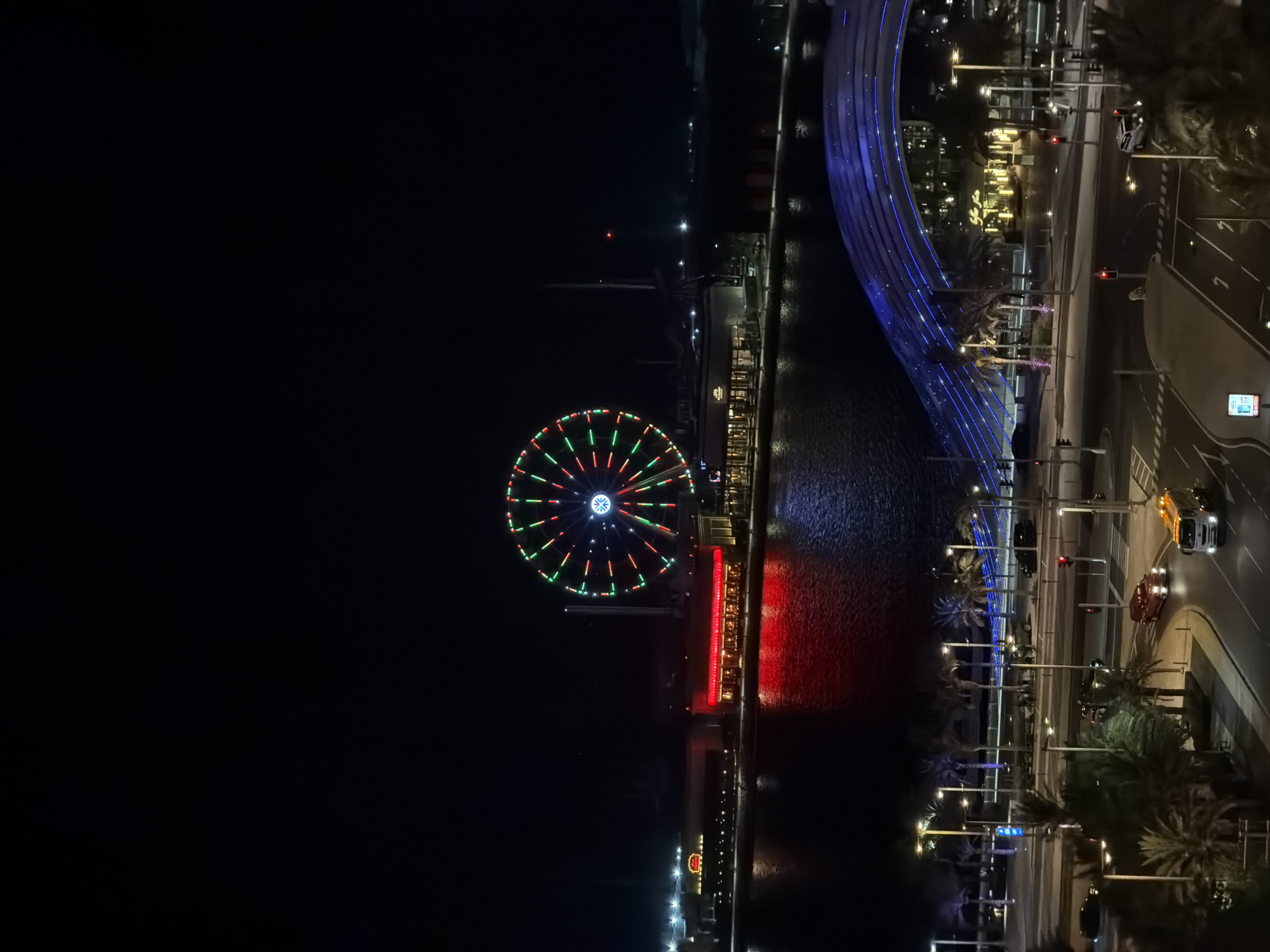 This image captures an elevated nighttime view of Bluewaters Island in Dubai, United Arab Emirates. The prominent feature is the Ain Dubai observation wheel, centrally located and illuminated with alternating red and green lights, featuring a blue and white light pattern at its hub. Directly below the observation wheel, along a dark body of water reflecting various lights, are several waterfront buildings with illuminated facades, including one with a red-lit exterior and another faintly displaying "The Cheesecake Factory" sign. To the right, a distinctive pedestrian bridge arches over the water, illuminated with multiple blue linear lights.
In the foreground, a multi-lane asphalt road is visible, bordered by sidewalks and numerous streetlights. Several vehicles are present on the road: a red classic-style car driving towards the lower-left, a white service truck with amber warning lights also moving towards the lower-left, and a white SUV stopped at a red traffic light on the right side of the road. Multiple traffic lights are visible, with most displaying red signals. Palm trees are scattered throughout the scene, lining the roads and waterfront. Small groups of indistinct people can be observed walking along the waterfront promenade and near the base of the blue-lit bridge. The sky is dark and clear, indicating nighttime conditions.
