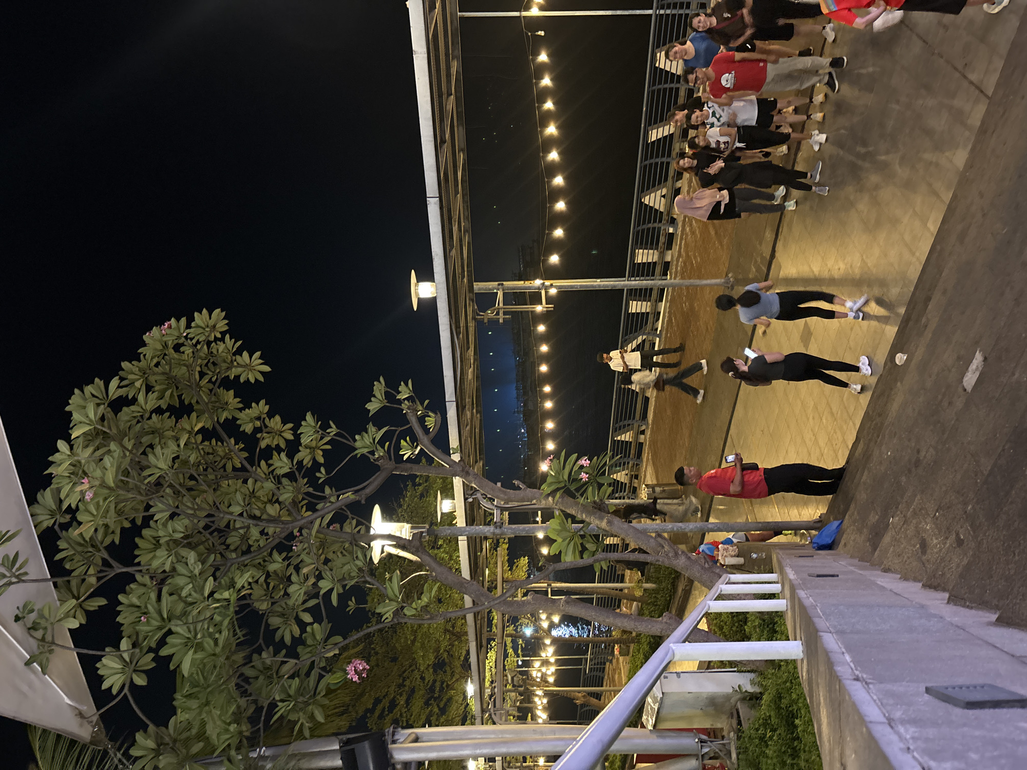 The image captures a nighttime scene along a waterfront promenade in North Jakarta, Indonesia. A group of people, appearing to be a mix of locals and tourists, are gathered on a paved walkway. In the foreground, a concrete staircase leads down towards the walkway. A man in a red shirt and black pants stands on the walkway, holding a phone. Further down the walkway, a group of women and men are conversing and standing. To the left, a tree with green leaves and pink flowers is visible, with lights strung along a railing in the background. The promenade overlooks a body of water, with distant lights reflecting on its surface. The overall atmosphere suggests a casual evening gathering in a public space.