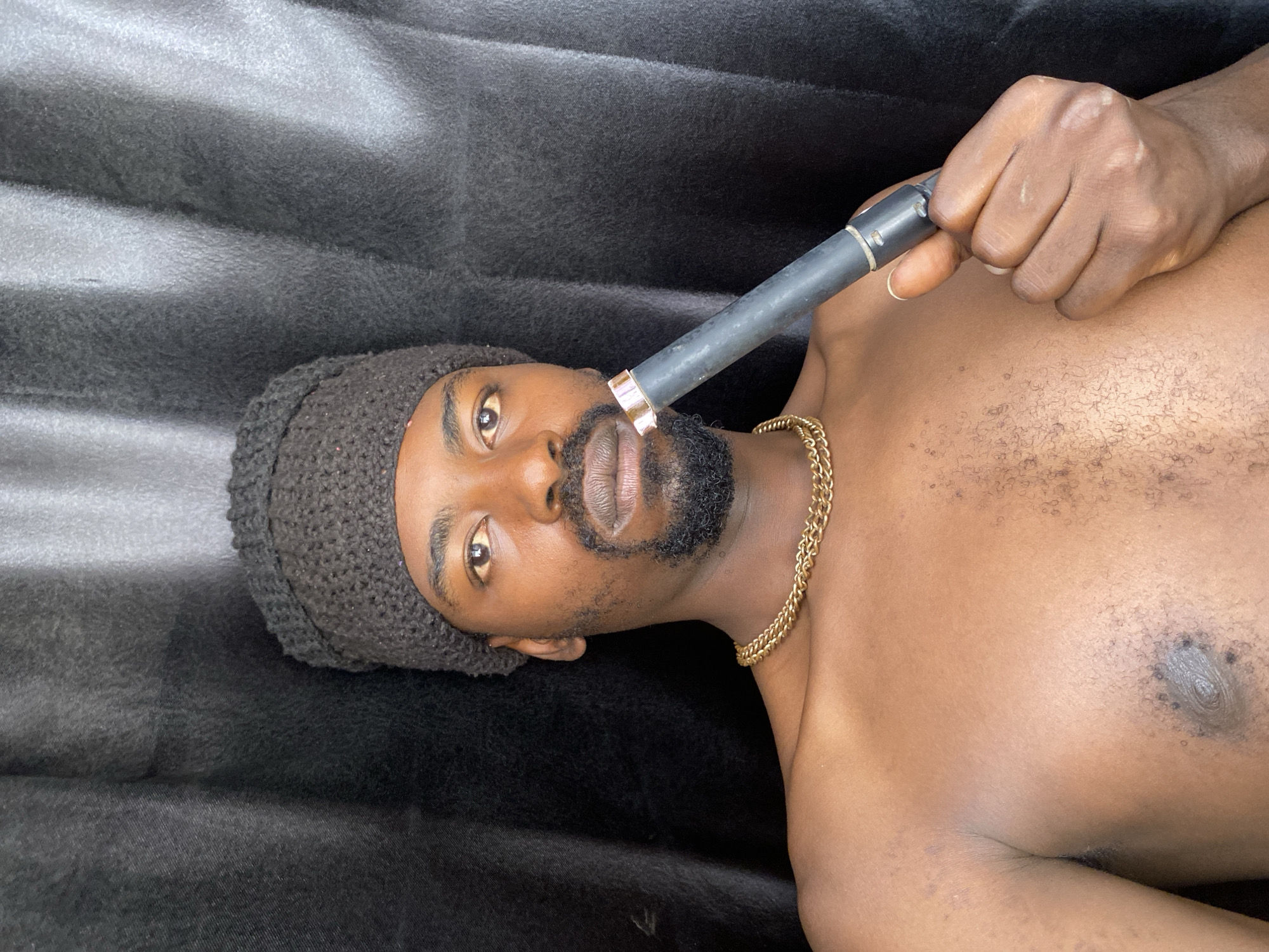 An adult male with dark skin and a beard is depicted in a medium close-up, lying on his back against a dark, textured fabric background. He is wearing a dark, knitted cap and a gold chain around his neck, and appears shirtless. He holds a black and gold tubular object, resembling a vape pen or electronic cigarette, to his mouth with his right hand. His eyes are open, and he looks directly into the camera with a neutral expression. The image is oriented with the man lying horizontally, so his head is on the left side of the frame and his body extends towards the right. His bare chest and right arm are visible, showing some body hair. The background is a solid, dark, soft material, possibly a blanket or bed covering. There is no visible text in the image. The scene takes place indoors.