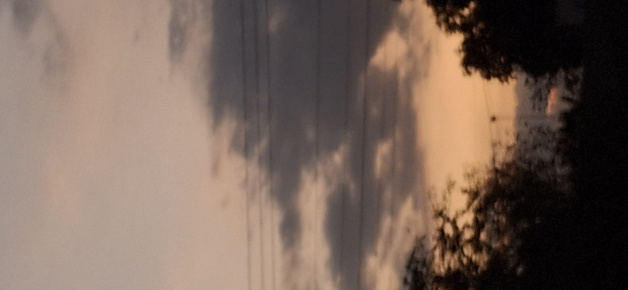 The image shows a partially cloudy sky at dusk or dawn, with colors ranging from light orange to gray. Power lines are visible against the sky. A dark, indistinct mass, possibly trees or foliage, is present on the right side. It is not possible to confirm if the photo was taken at Vallecito, Venezuela from this image. There are no people visible in the image.