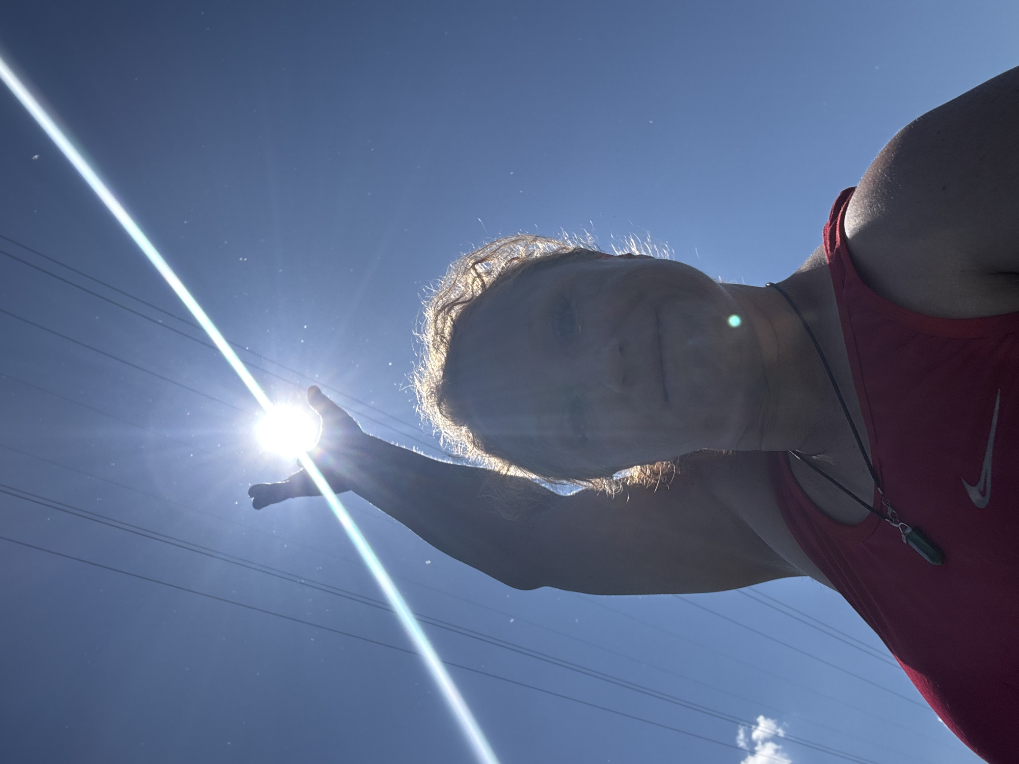 A low-angle, outdoor, eye-level, medium shot captures a person wearing a red tank top with a Nike logo and a dark necklace with a green pendant, silhouetted against a bright blue sky. The sun is visible in the upper center of the frame, with a prominent lens flare streaking across the image. The person's right arm is raised, with their hand positioned to obscure the sun. The person has light-colored, curly hair, and their face is partially visible, looking upwards. Three parallel power lines stretch diagonally across the sky from the upper left to the upper right. The scene appears to be a sunny day. The location is not discernible from the image.