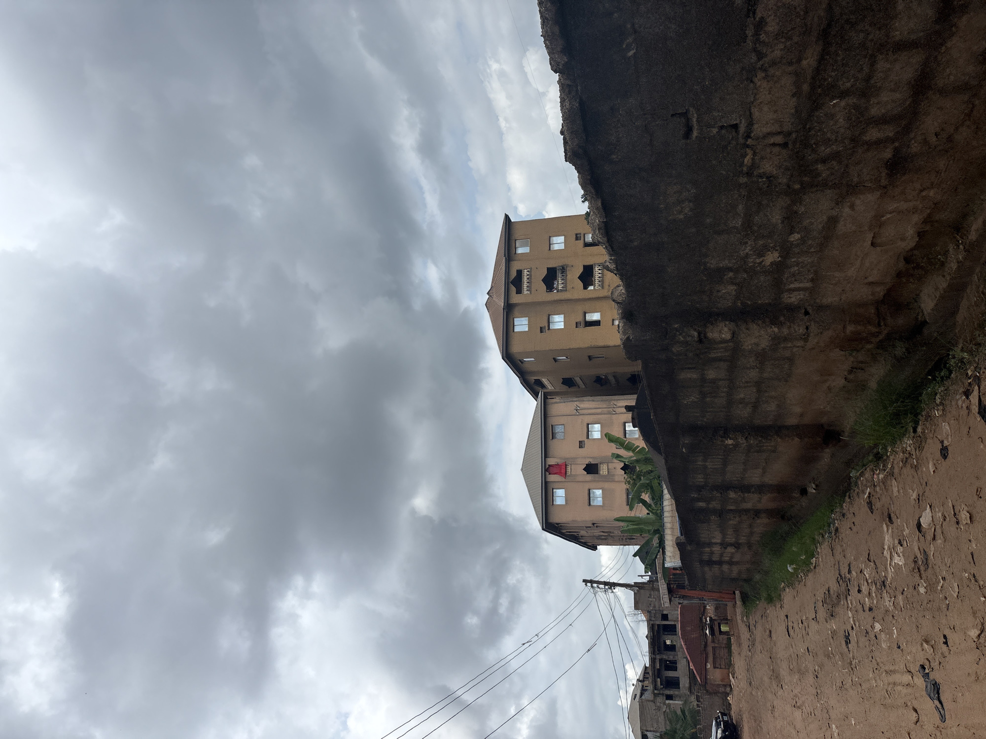 The image depicts a street scene in Nkpor, Nigeria, under a cloudy sky. In the foreground, a dirt road with scattered debris leads past a rough, dark stone wall. Beyond the wall, two multi-story buildings with tan facades stand prominently. The building on the right has balconies and multiple windows, while the one on the left features a red element on its roofline and several windows. Utility lines and poles are visible to the left of the buildings, and other structures, including unfinished or partially constructed buildings, are present in the background. Banana plants are growing near the buildings. The overall impression is that of an urban environment with signs of ongoing development or construction.