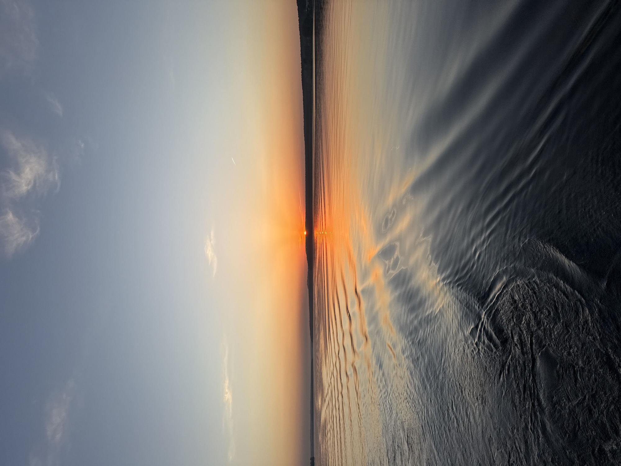 The image shows a sunset over a body of water, likely a lake or river, in Votkinsk, Russia. The sun is low on the horizon, creating a bright orange glow that is reflected in the water. The sky is a mix of blue and orange hues, with a few scattered clouds. The water is relatively calm, with gentle ripples on the surface. On the horizon line, there is a treeline visible. There are no people visible in the image.